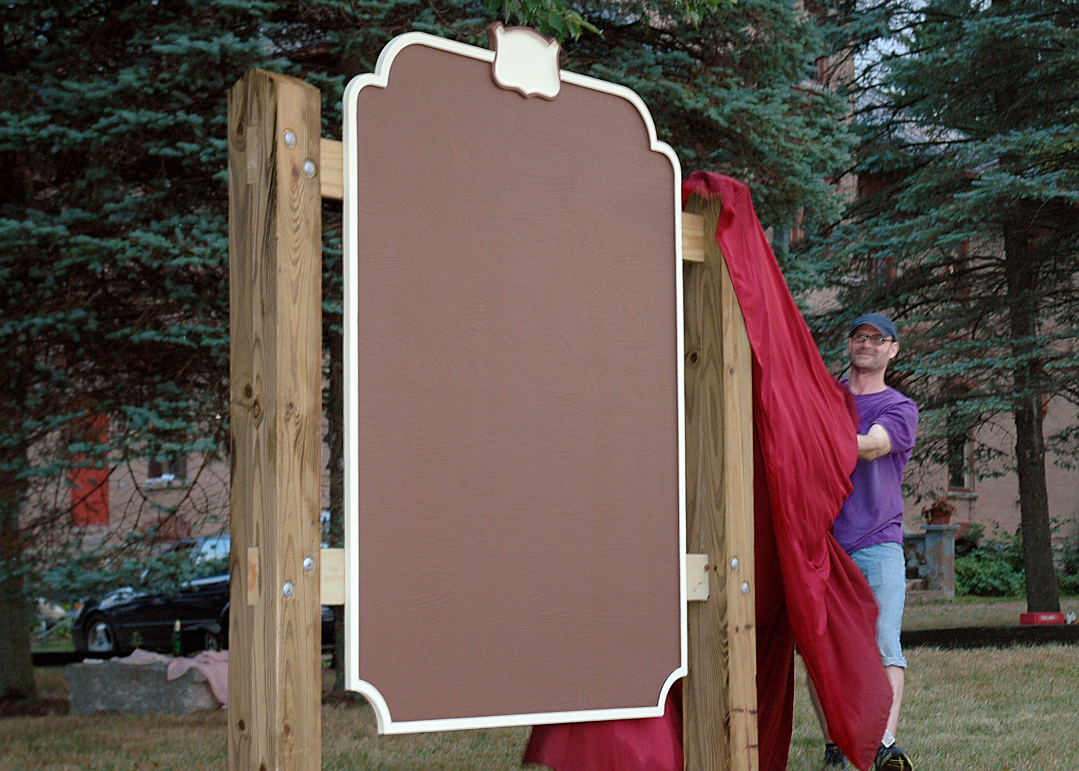  Man in purple shirt, jean shorts, and black hat fully removes red fabric, revealing plaque.