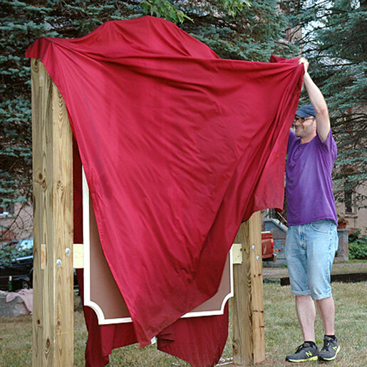 Man in purple shirt, jean shorts, and black hat begins to pull back red fabric, revealing plaque.