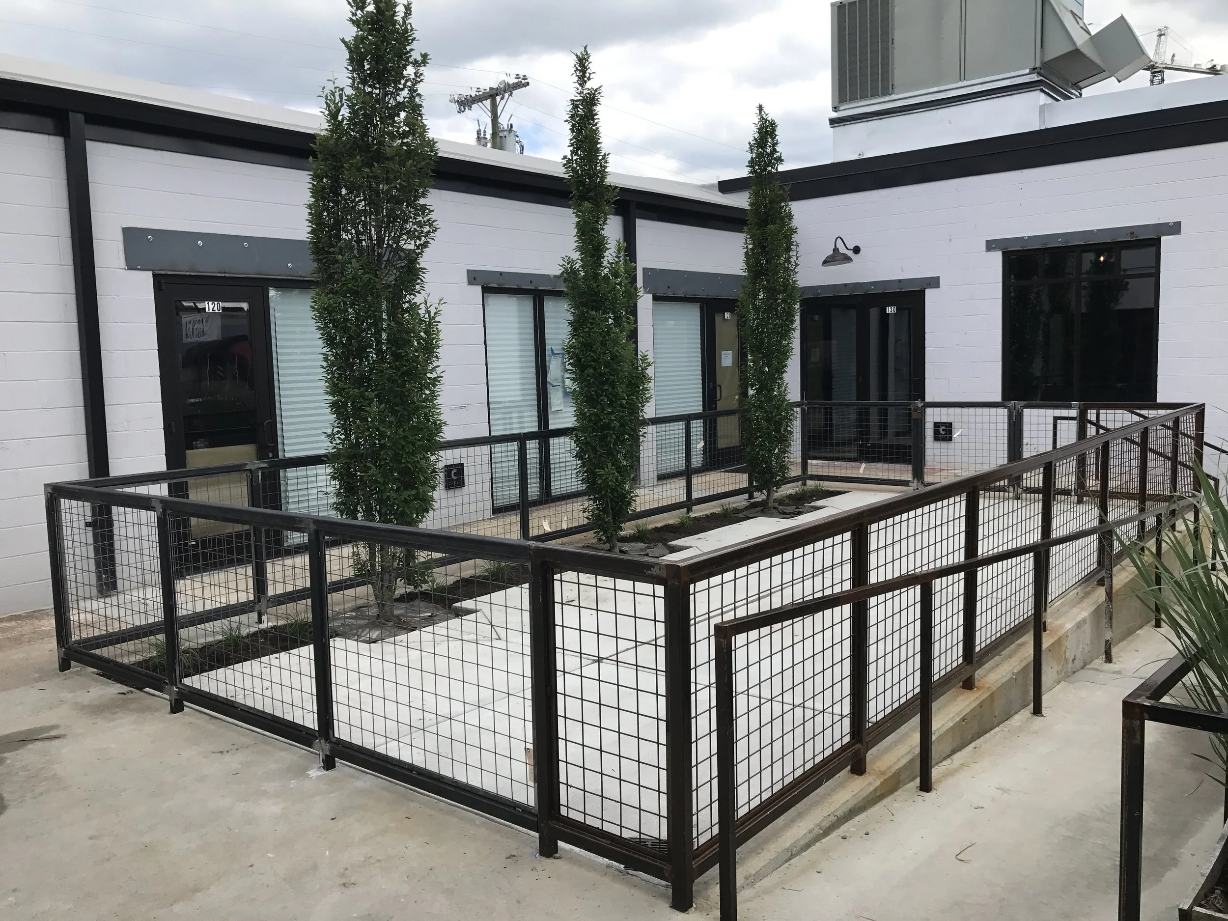 Outdoor area with small trees, black metal fencing, white brick building with windows and doors, and outdoor lighting fixtures.