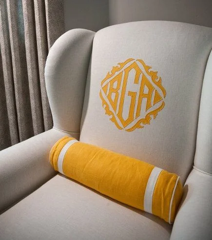 A wingback chair upholstered in linen with an embellished  monogram and coordinating bolster