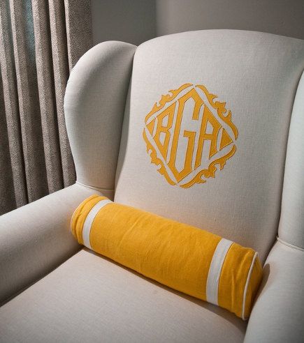 A wingback chair upholstered in linen with an embellished monogram and coordinating bolster