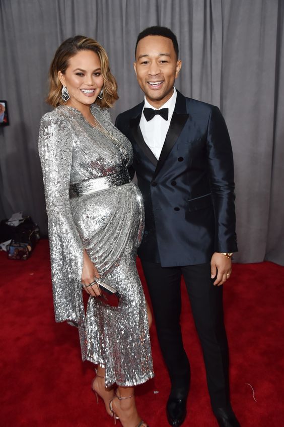 Chrissy Teigen is glowing in a Yanina Couture dress that accents her growing baby (boy) bump. She's also wearing lorraine schwartz jewelry. The always smiling john legend is wearing a Burberry suit, chopard jewelry and bruno magli shoes. 