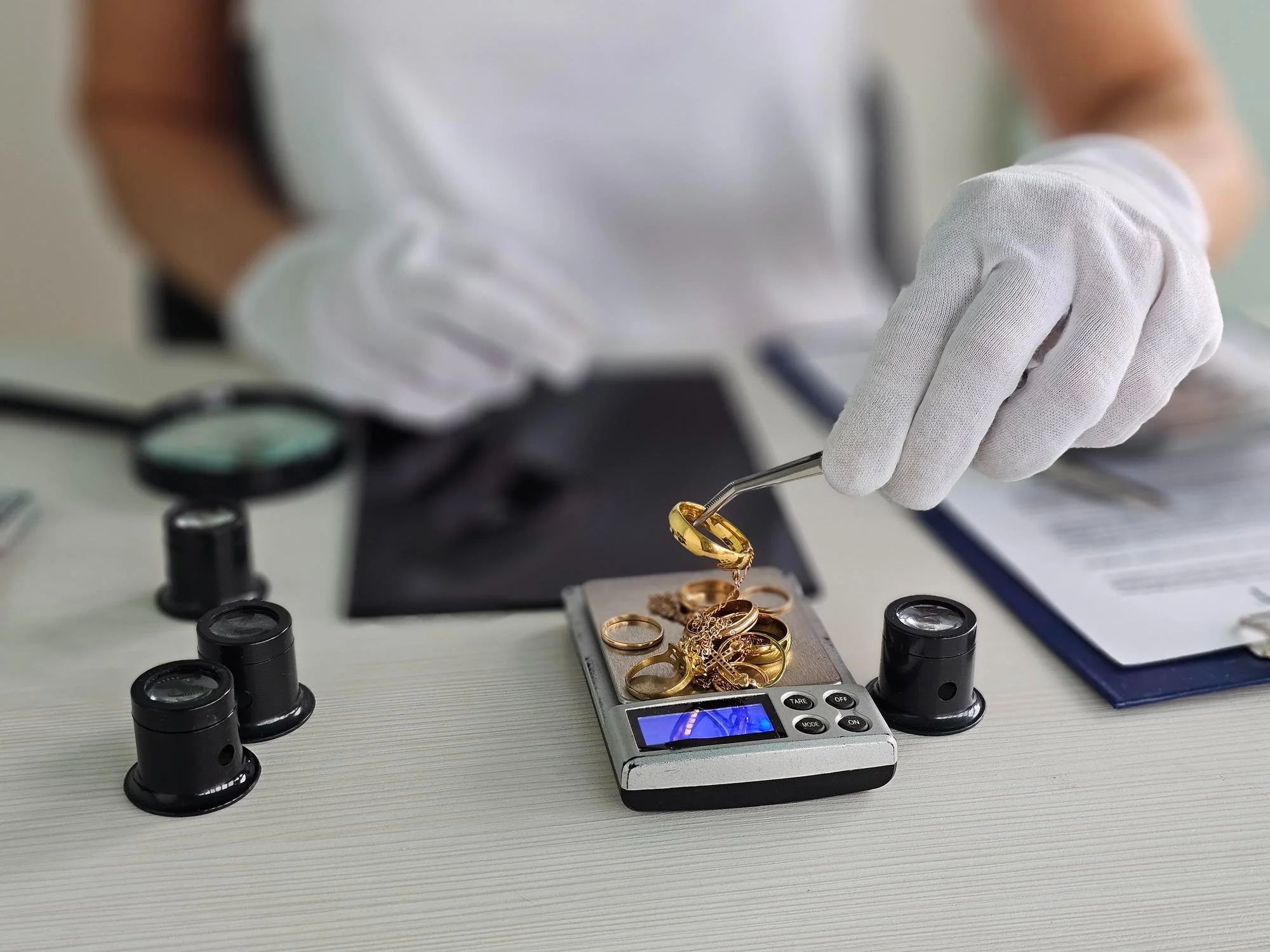 Close-up of a person's hand wearing a white glove using tweezers to handle a gold ring on a digital scale. The scene includes several other gold rings and jewelry pieces on a gray surface, with a magnifying glass and other tools scattered around.