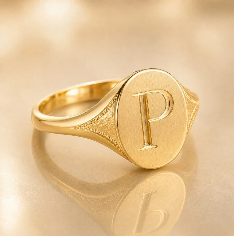 A gold signet ring with a large round face engraved with the letter 'P'. The ring is reflective and placed on a smooth surface.