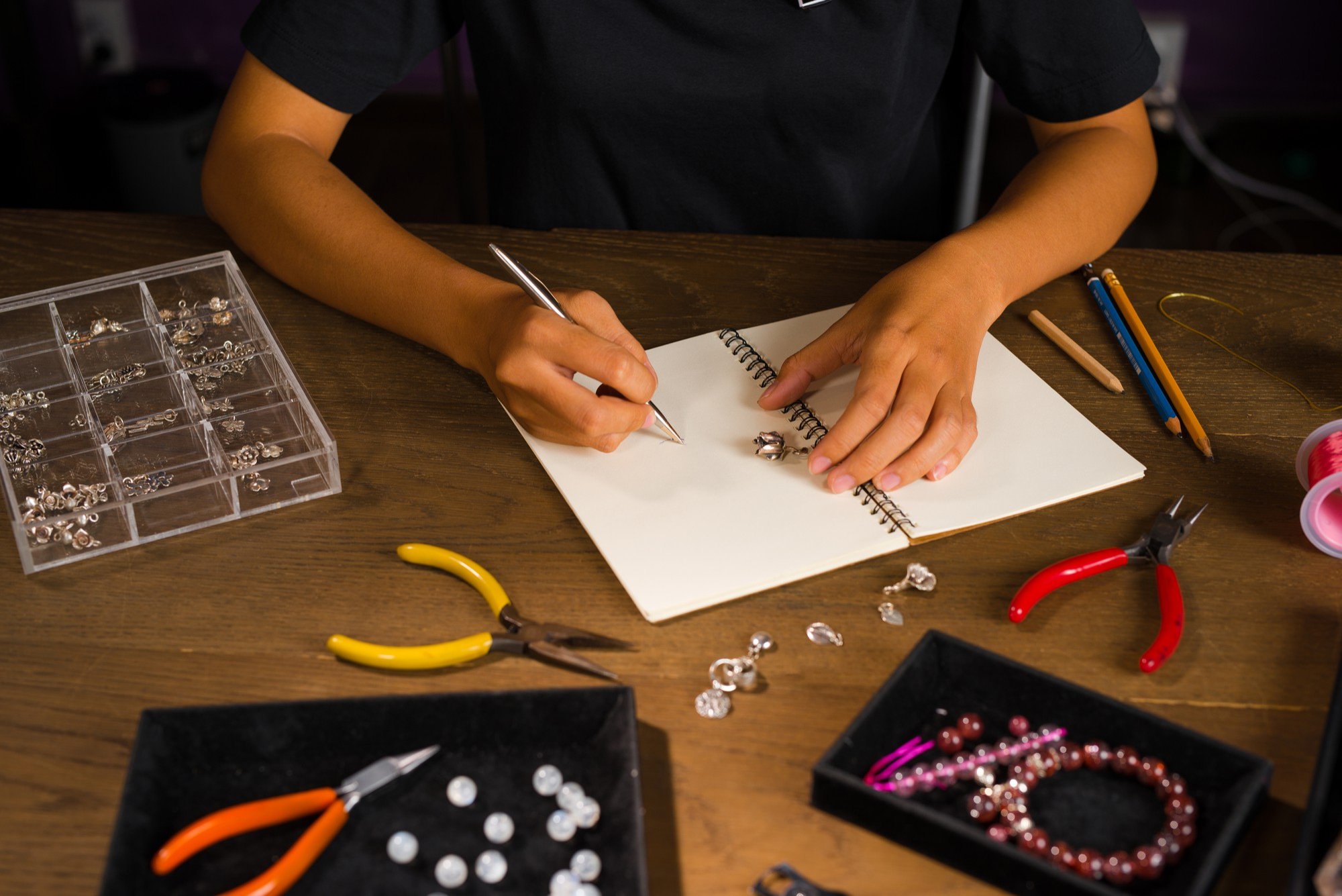 Person working at a jewelry-making station, with jewelry pieces, pliers, and tools on a wooden table.