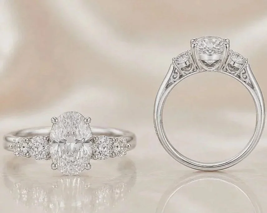 Two diamond engagement rings, one with an oval center stone and side stones on a silver band, the other with a round center stone and side stones on a silver band, displayed on a reflective surface.