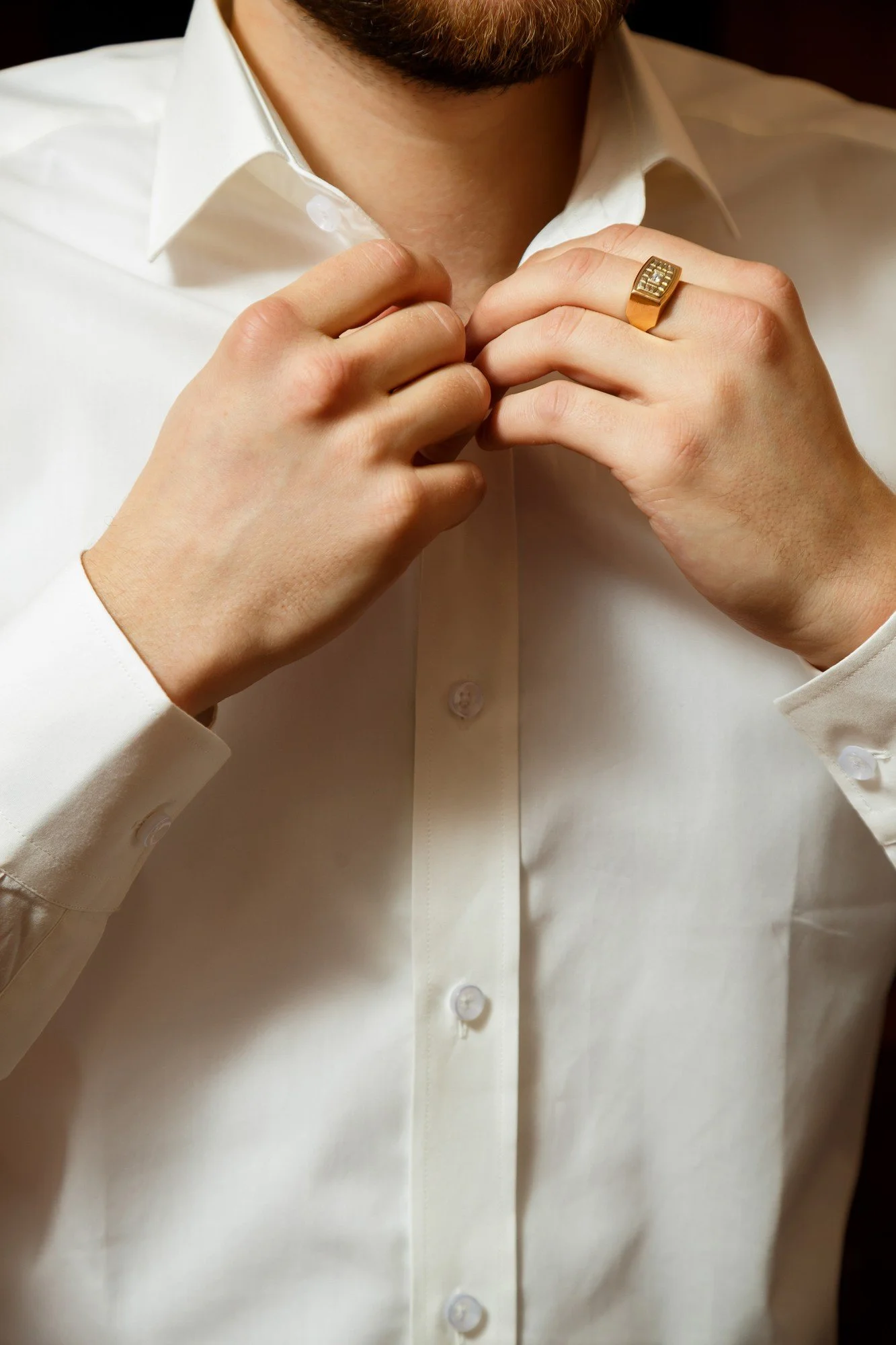 A man adjusting a white dress shirt with both hands, wearing a gold ring with diamonds on his right hand.