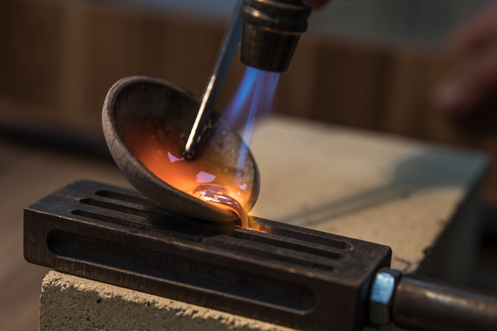 A small, metal pipe is being heated with a blowtorch, causing a bright orange glow inside it. The blowtorch’s blue flame is directed at the pipe, which is placed on a black surface with slits.