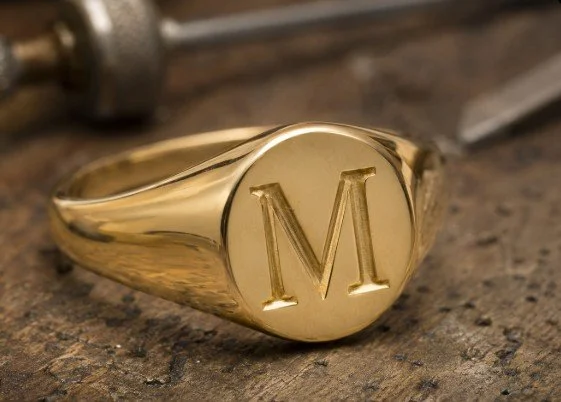 Close-up of a gold signet ring with the letter 'M' engraved on it, resting on a textured surface.