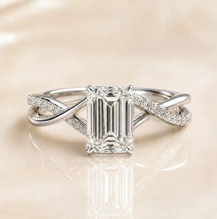 Silver ring with a large rectangular emerald-cut diamond center stone, accented by smaller pave-set diamonds on the band, placed on a reflective light-colored surface.