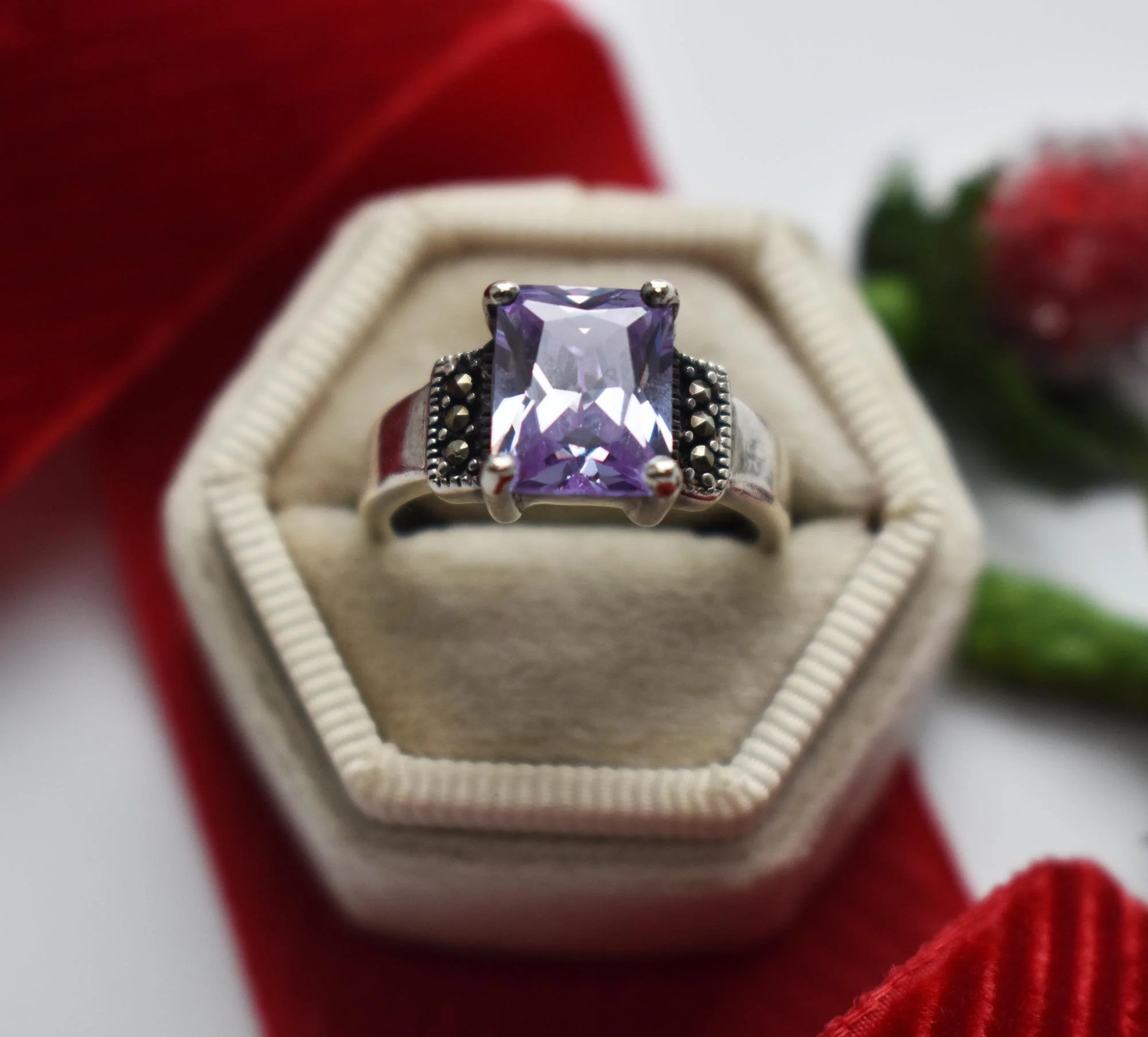Close-up of a silver ring with a large rectangular purple gemstone. The ring is displayed inside a beige ring box, surrounded by a red cloth and green foliage in the background.