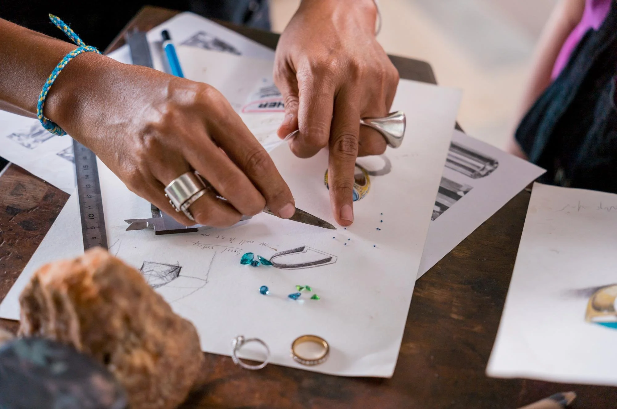 Hands of two people working on jewelry designs on paper, with jewelry tools, rings, and decorative stones on the table.