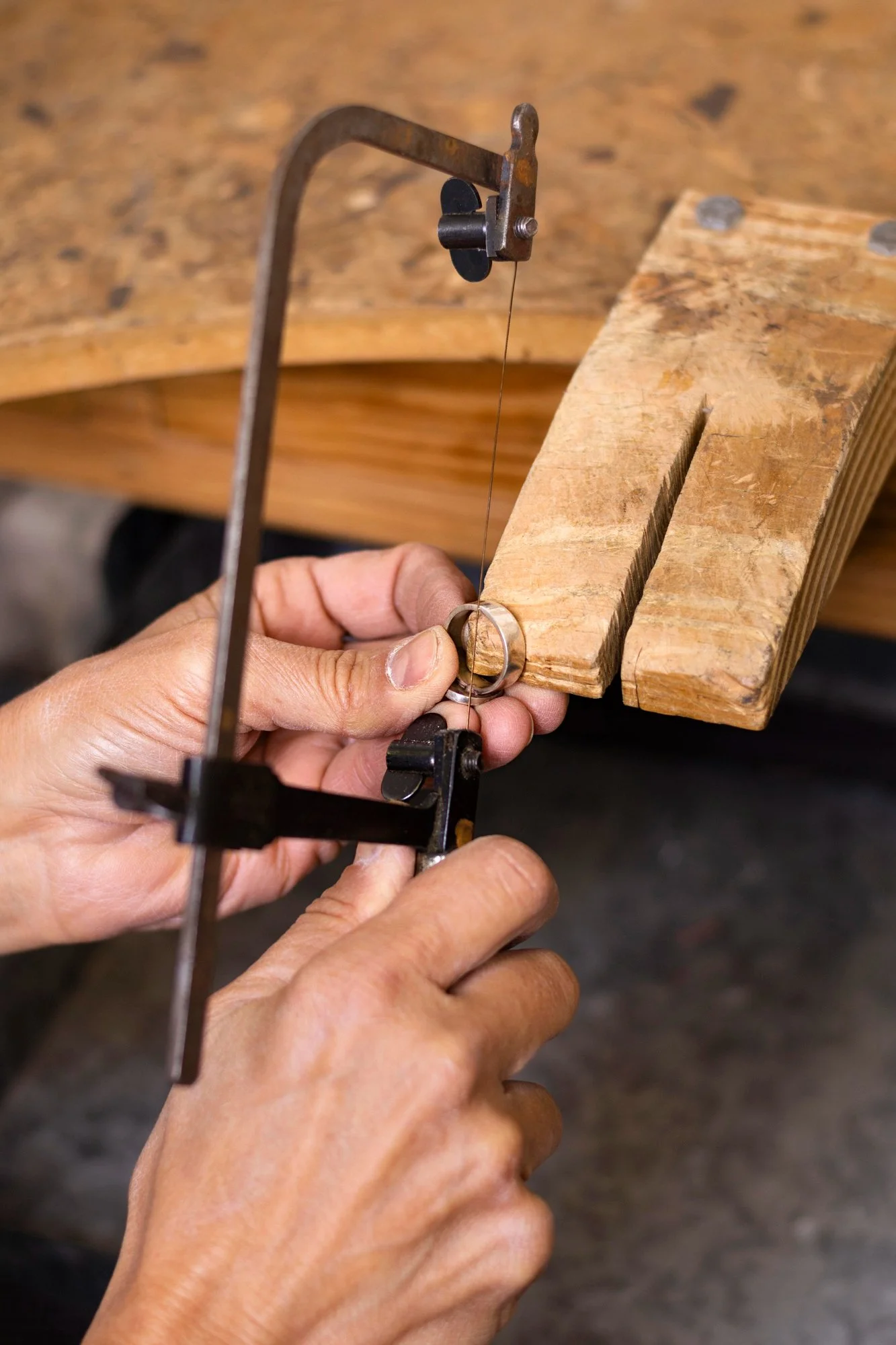 Close-up of a person's hands using a coping saw to cut a metal ring, with a piece of wood clamped as a work surface.