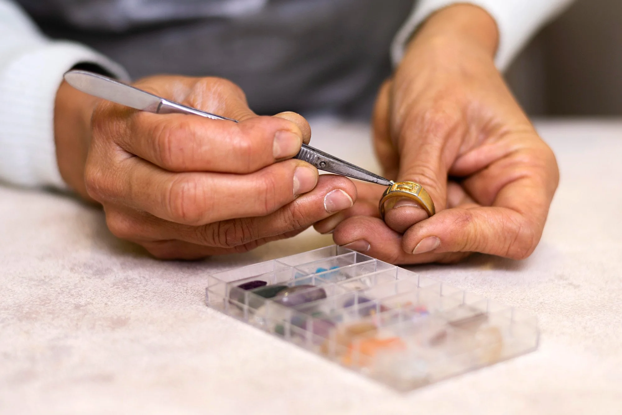 Close-up of a person's hand using a manicure tool on a gold ring, with a plastic container of colorful gemstones in the background.