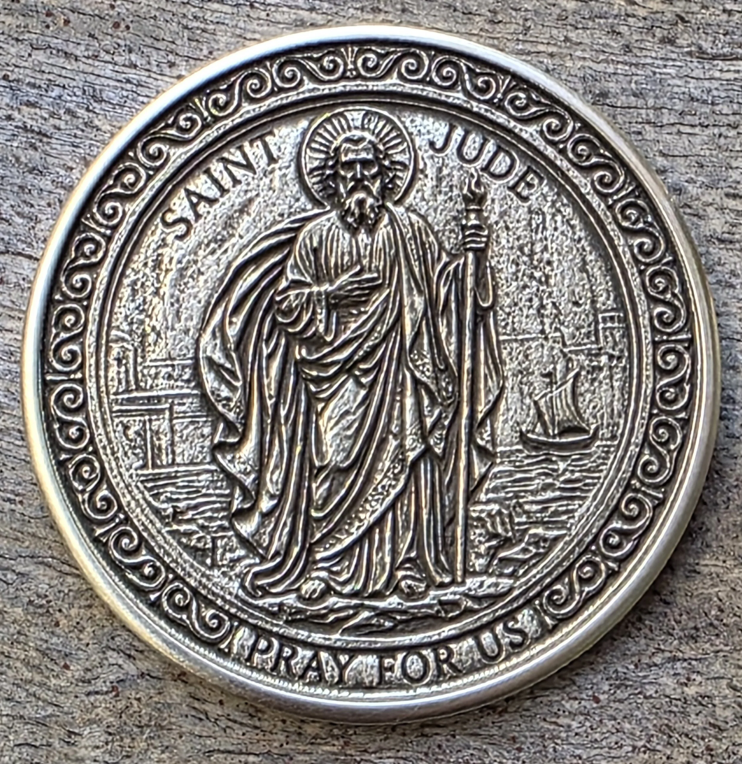 Close-up of a silver coin depicting a religious figure in the center. The coin has intricate border designs and text around the edge that reads 'Saint Jude' and other partially visible letters.