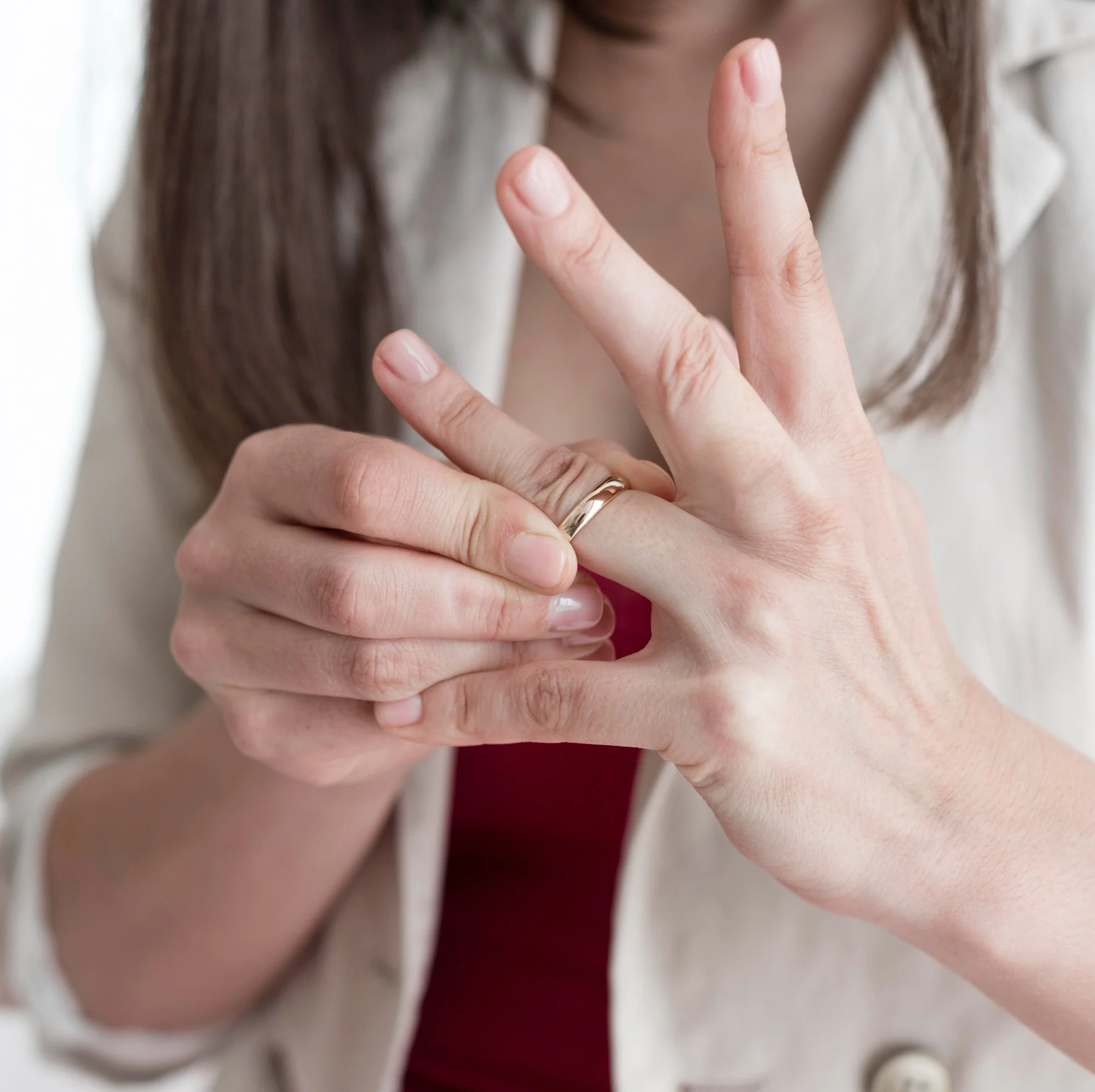 Close-up of a woman with light skin making a peace sign with her fingers, wearing a gold ring, and a white jacket over a red shirt, with her face partially out of focus in the background.