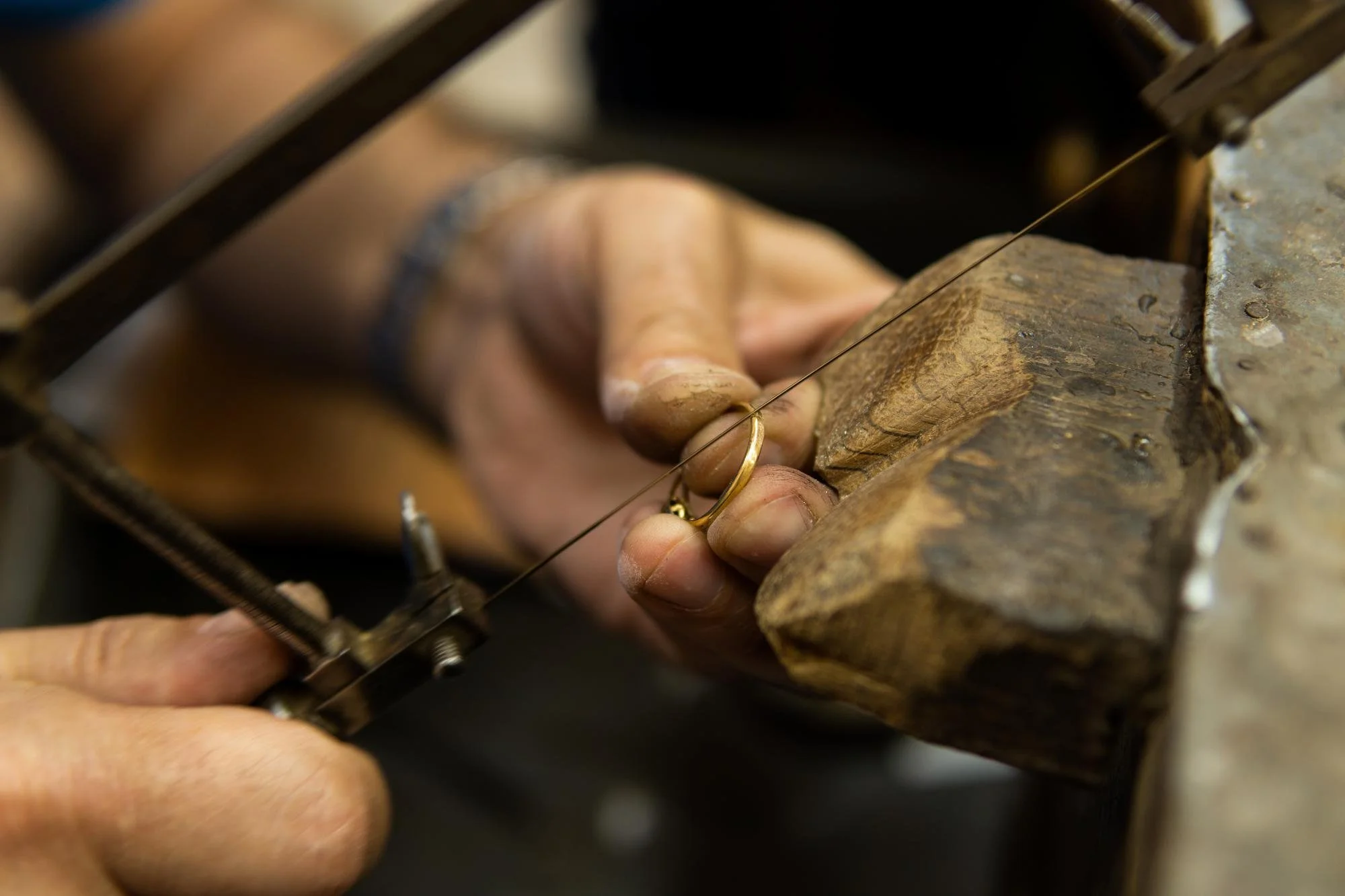 Close-up of a person using a small hand tool and a file to work on a metal jewelry piece in a workshop with a wooden workbench.