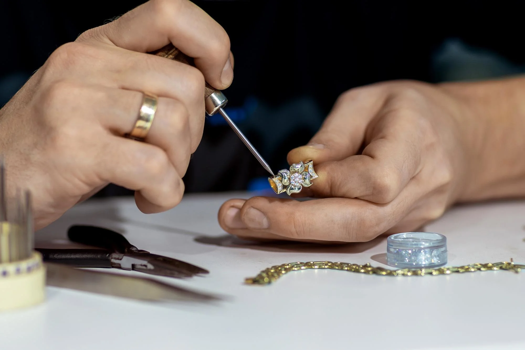 Close-up of a person using a tiny tool to work on a gold ring with diamonds, surrounded by jewelry and tools on a white surface.