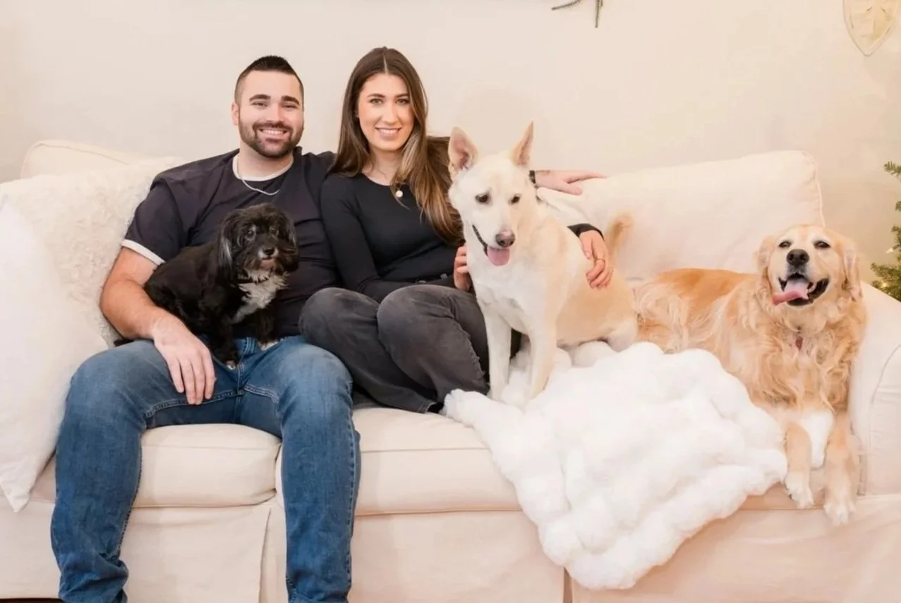 A couple sitting on a beige couch with three dogs, one small black, one medium-sized white with pointy ears, and one golden retriever, in a cozy living room.