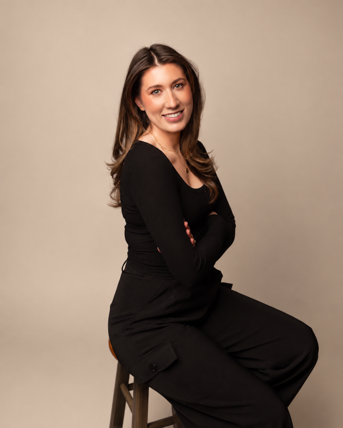 A young woman with long brown hair and a black long-sleeve top sitting on a stool against a plain background, smiling with crossed arms.