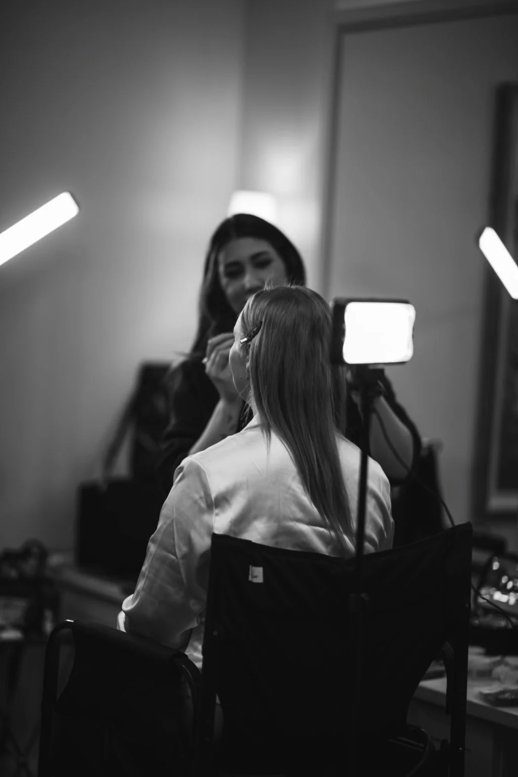 A woman is getting her makeup done by another woman near a mirror with a bright light. The scene is in a room with subdued lighting, and there is a smartphone on a selfie stick capturing the moment.