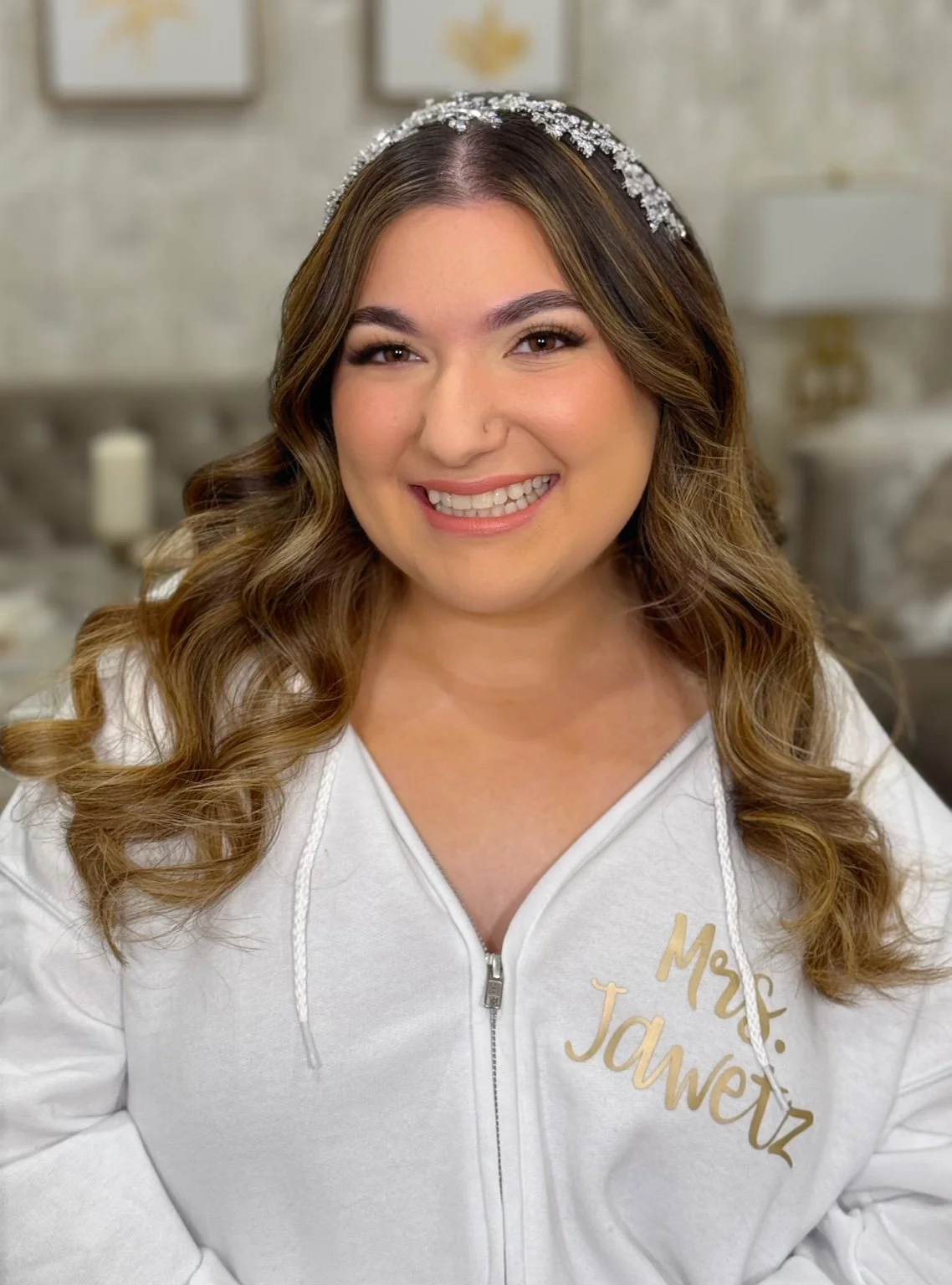 A smiling woman with long wavy brown hair wearing a white hoodie with gold text that says "Mrs. Jawetz" and a jeweled headband. She is in a cozy room with blurred background decor.