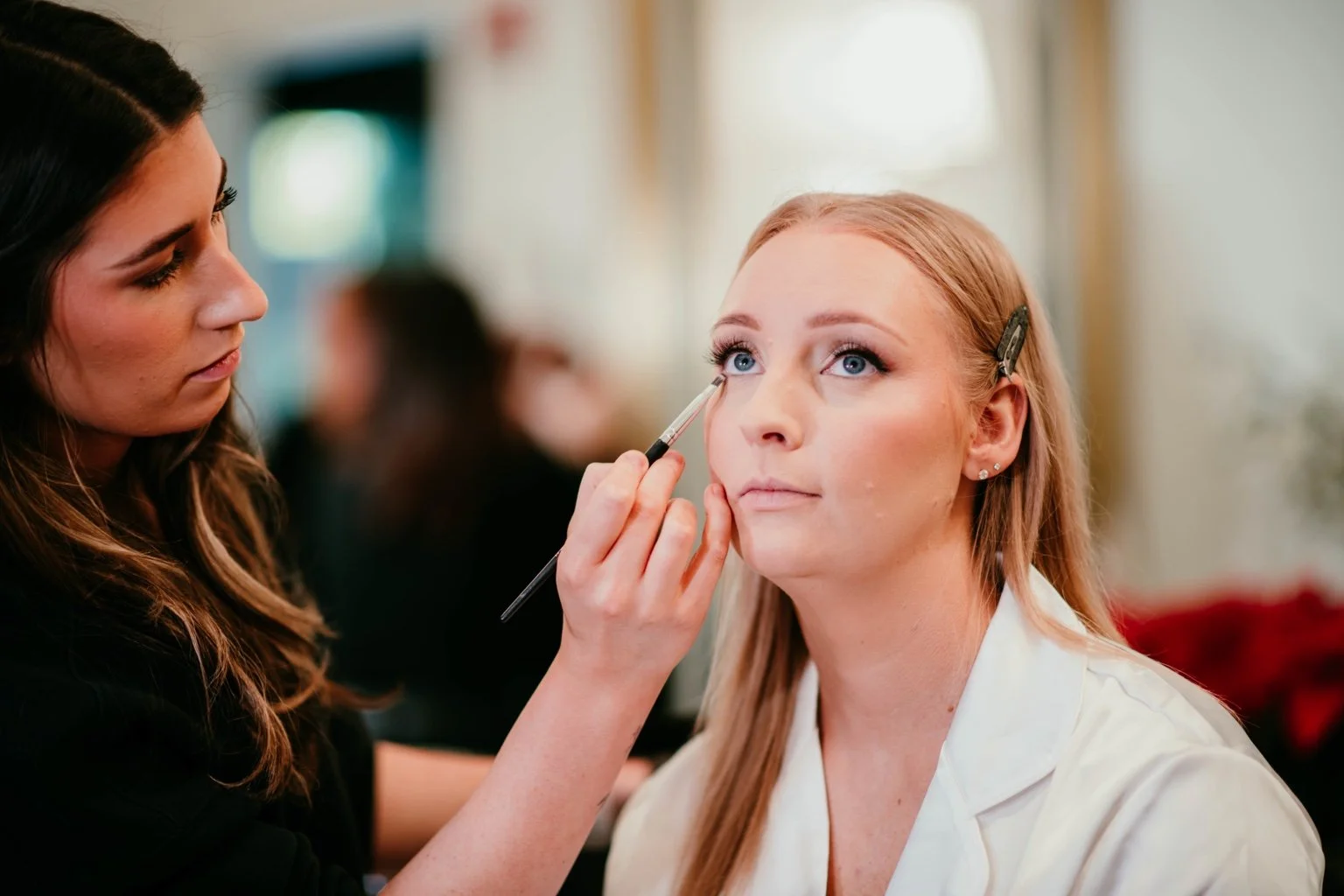 A woman with long, blonde hair and light skin has makeup applied by a makeup artist at a salon or studio. The makeup artist, with long, dark hair, is holding a brush near the woman's eye.