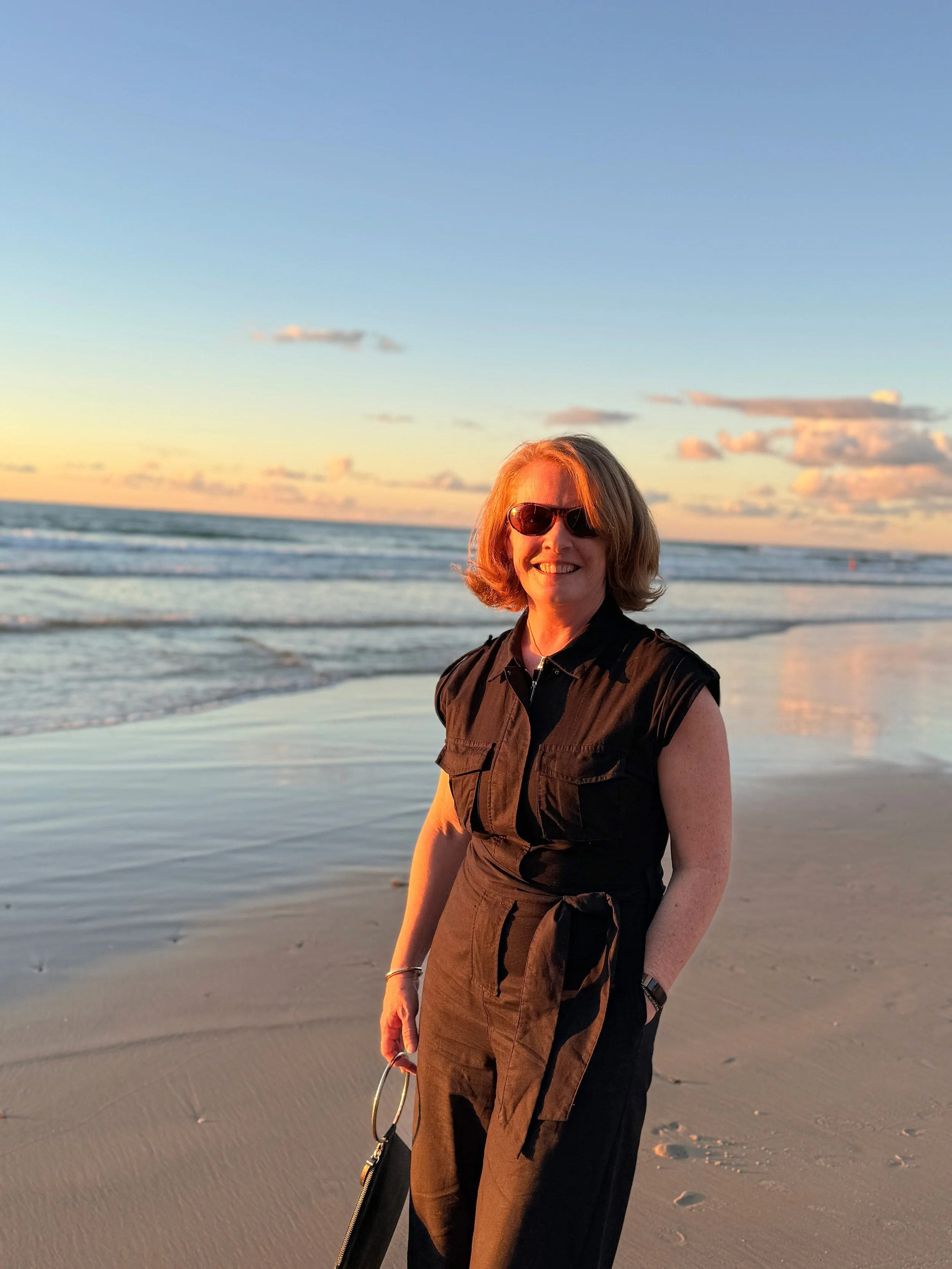 A woman with shoulder-length hair wearing sunglasses and a black sleeveless vest stands on a sandy beach during sunset, with the ocean and clouds in the background.