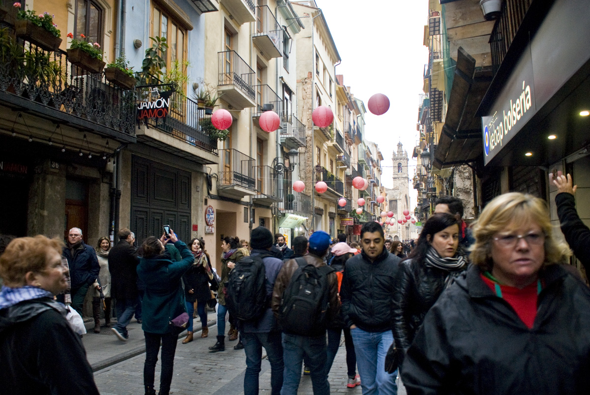 A street in the old part of the city, decked out for Fallas and filled with people celebrating.