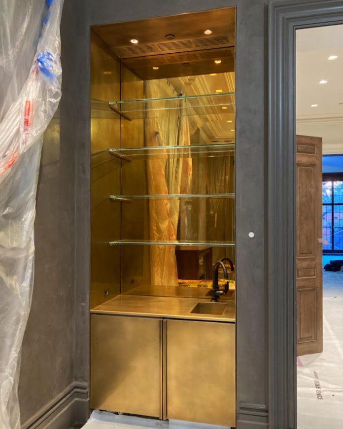  Custom inset hand patinated bronze bar, with seamless welded bronze sink, custom bronze cabinet and pulls with glass shelves in front of antiqued golden glass mirror. Fabricated for Kin and Company 
