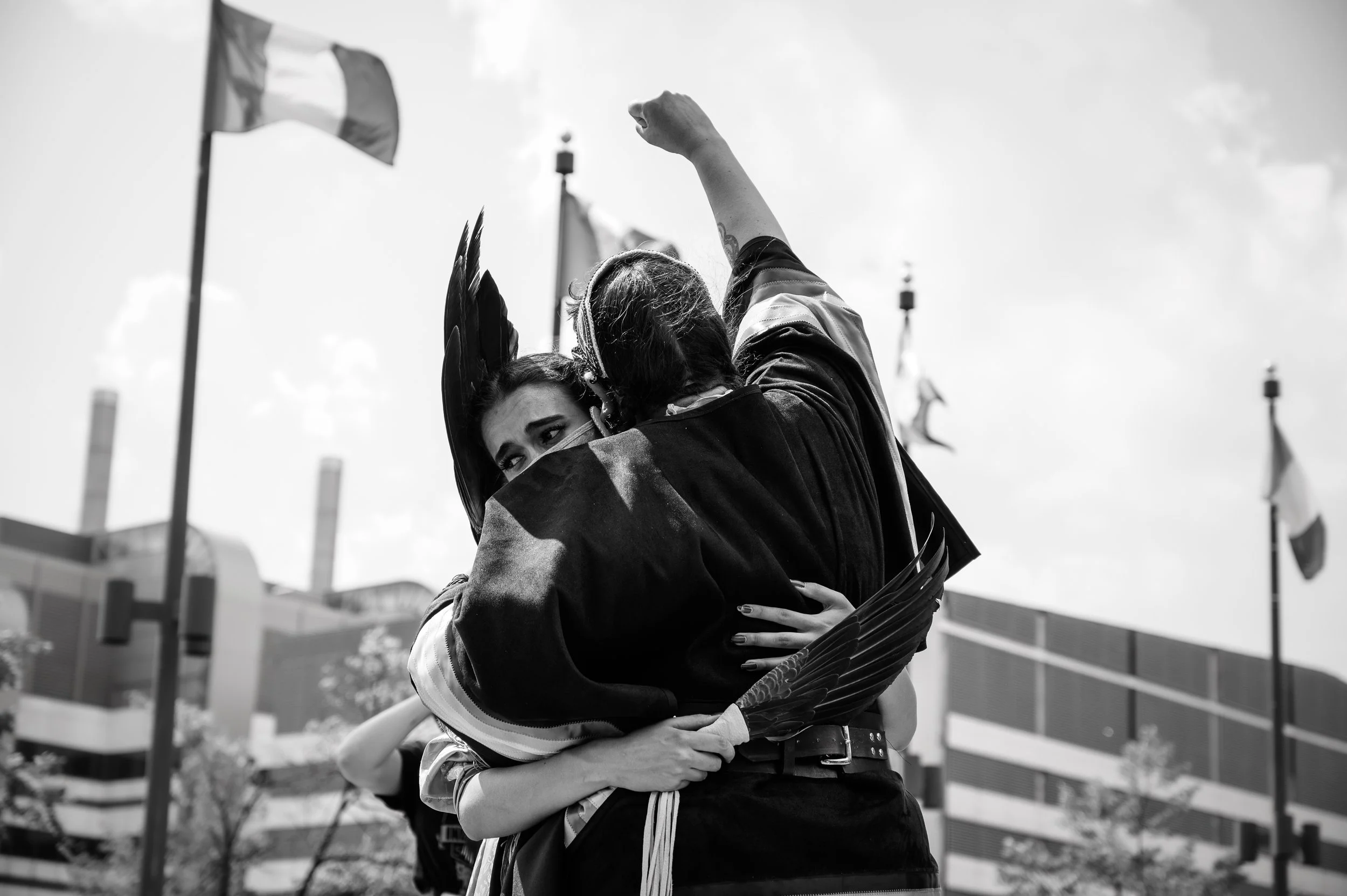 Two people hugging at an outdoor event, one with a patriotic flag draped over their shoulders, with flags flying in the background.