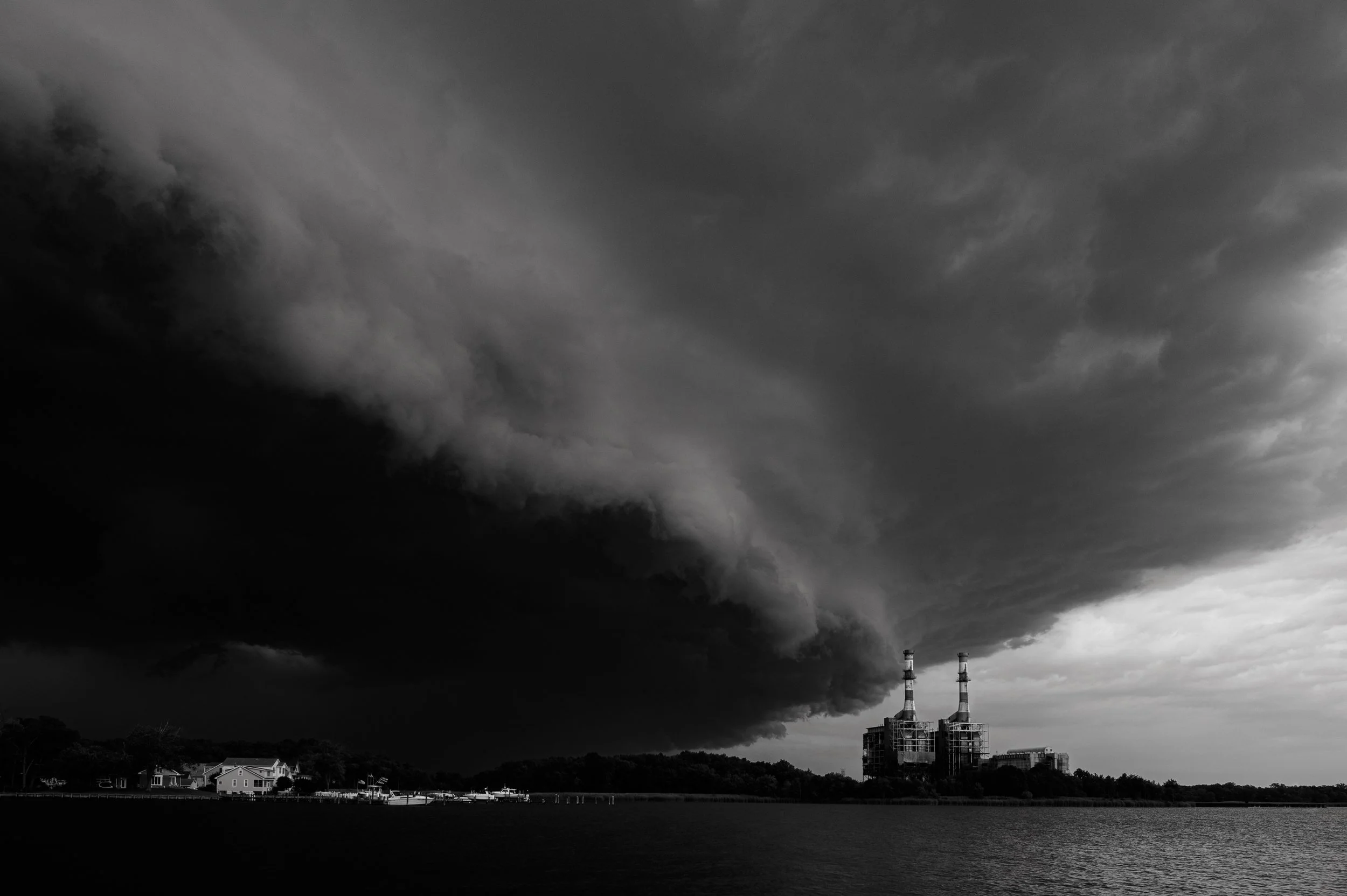 Dark storm clouds over a power plant with two smokestacks, a water body, and a cluster of houses in the foreground.