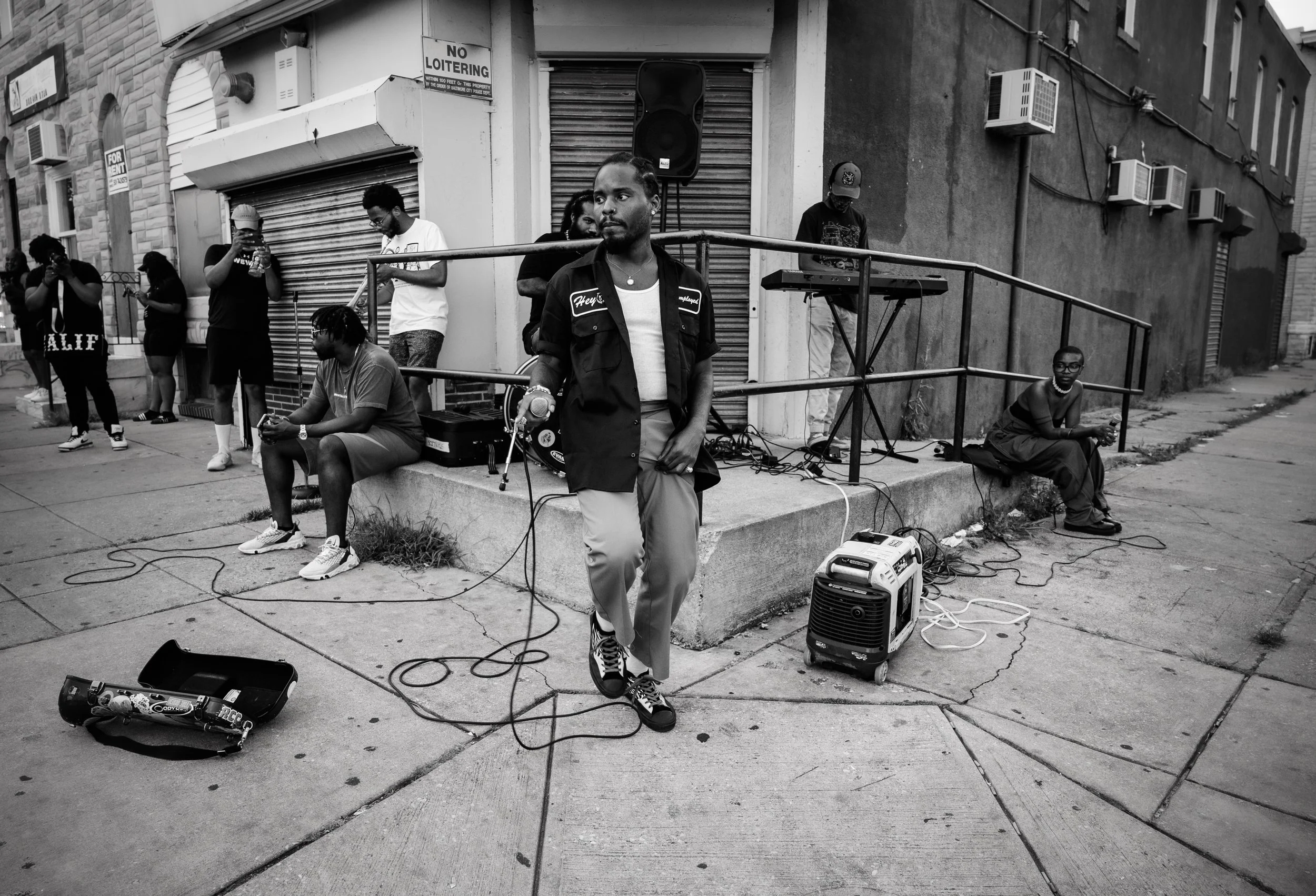 A man standing with a microphone in front of multiple people on a city sidewalk, with a portable speaker, pedal, and cables nearby, some people looking at their phones, others adjusting equipment, in a black-and-white photo.