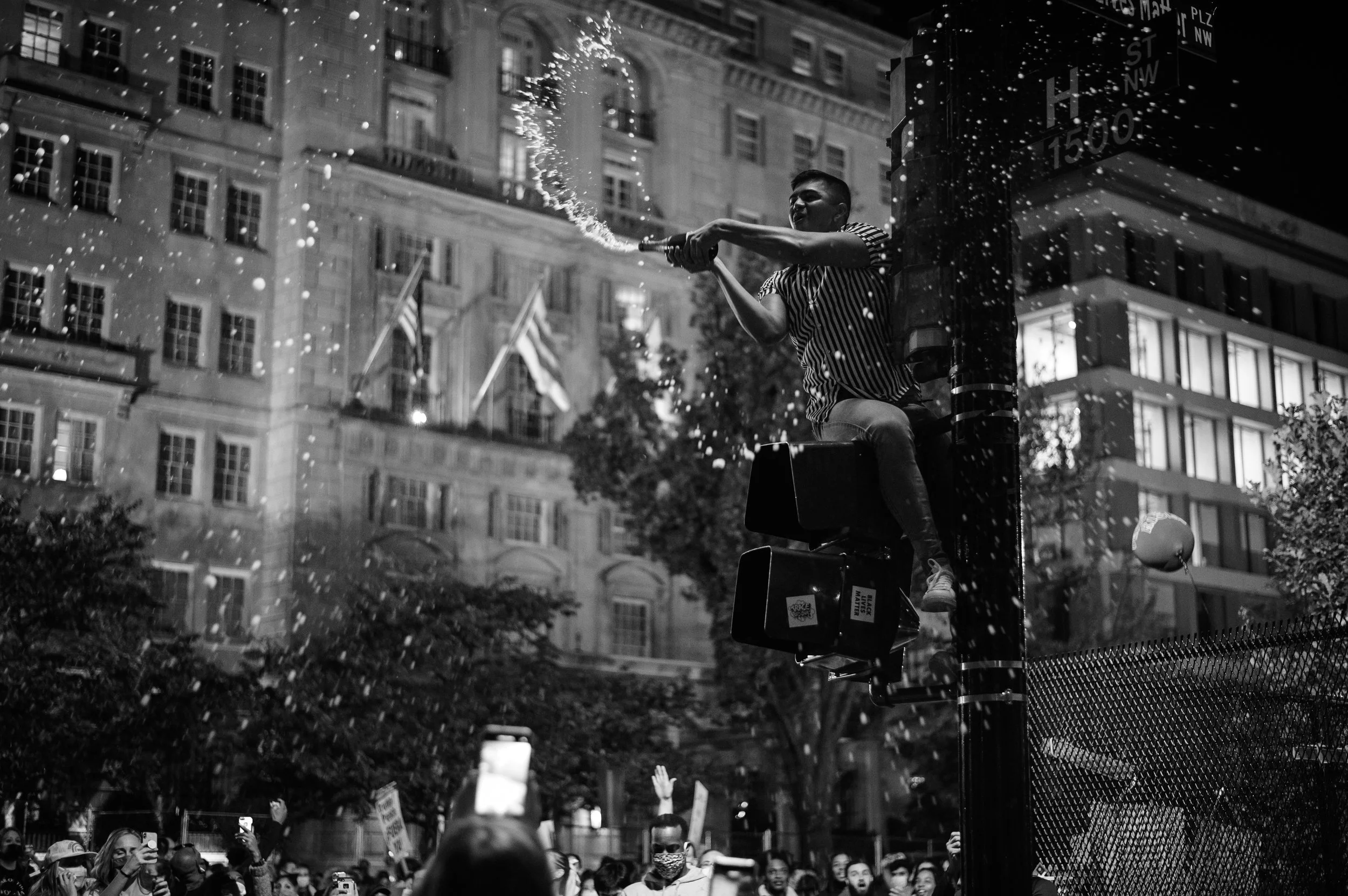A man in a striped shirt sits on top of a utility box, spraying water into the air with a hose during a night protest or demonstration in an urban area, surrounded by a crowd of people holding signs, with tall buildings in the background.