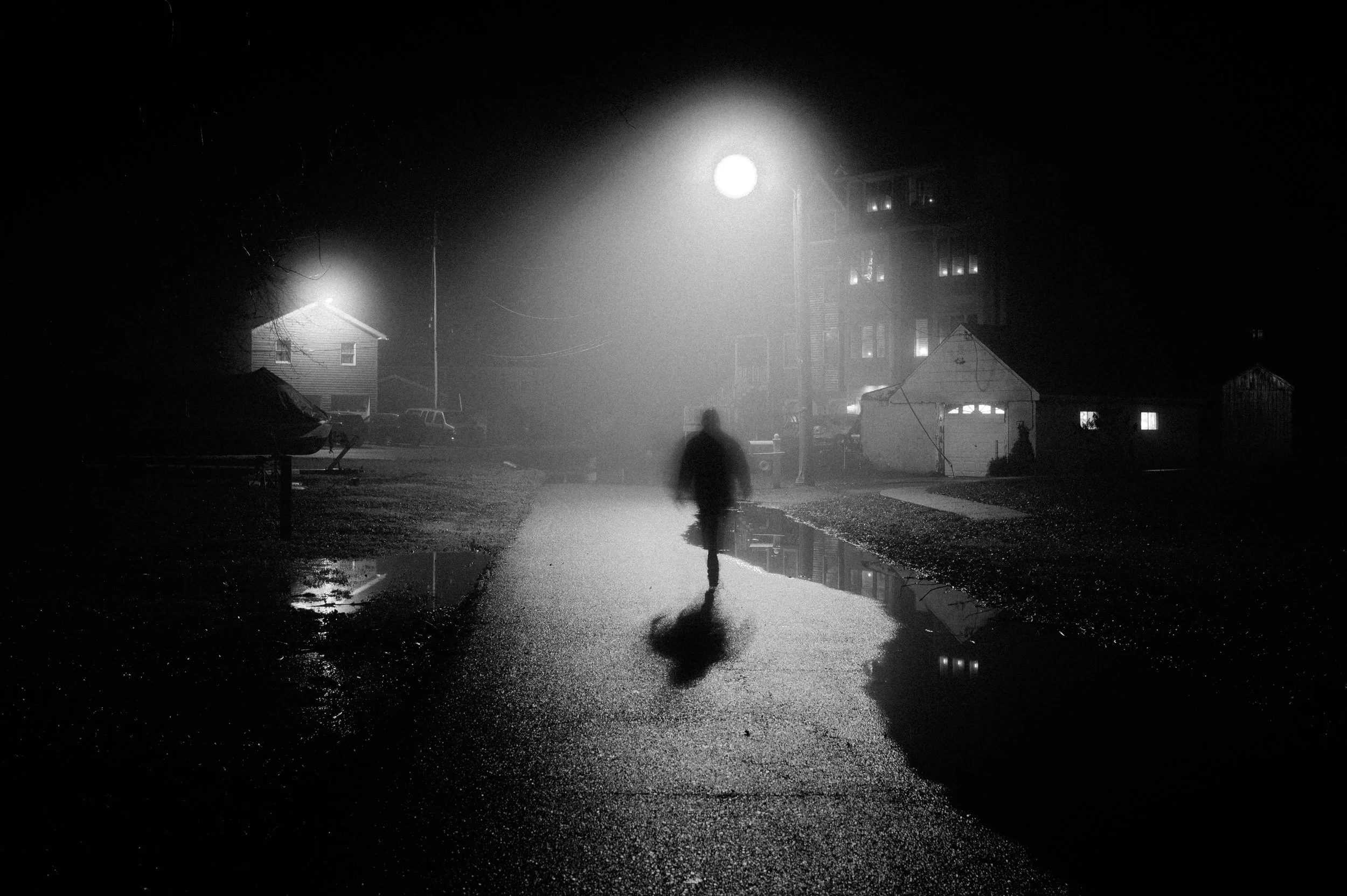 A black and white photo of a person walking on a wet street at night, with streetlights illuminating the scene and a large puddle reflecting the person and lights.