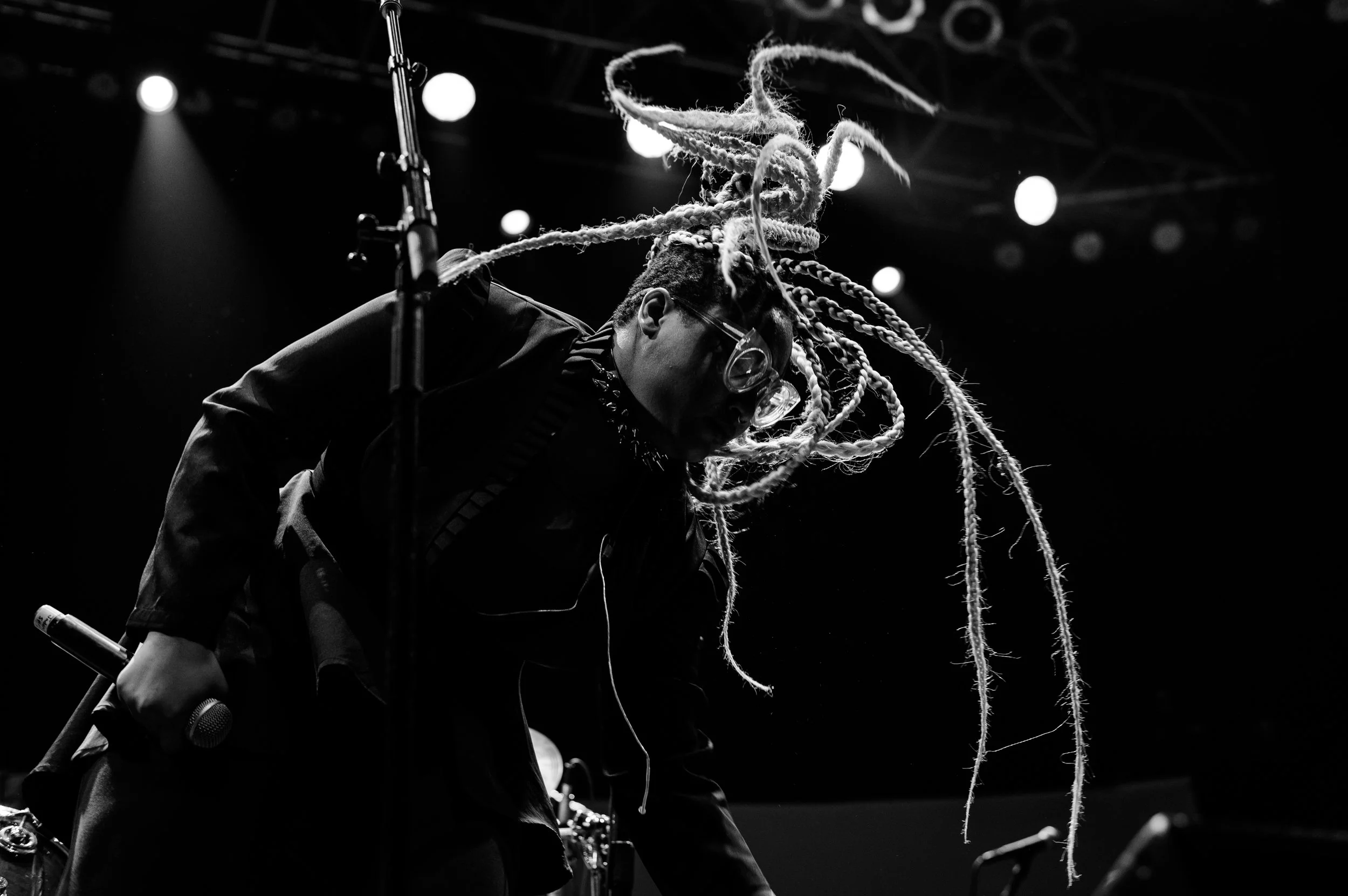A person performing on stage, with dreadlocks, wearing glasses, illuminated by stage lights, black and white photograph.