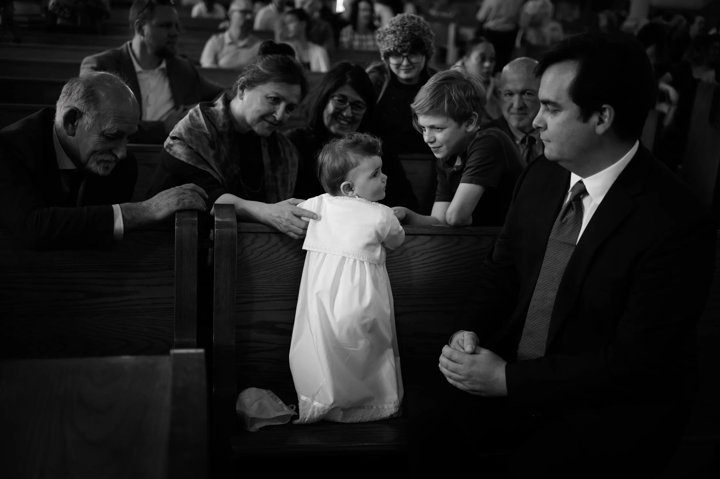A young girl in a white dress talking to a man in a suit, while other people watch, sitting in pews of a church or similar setting.