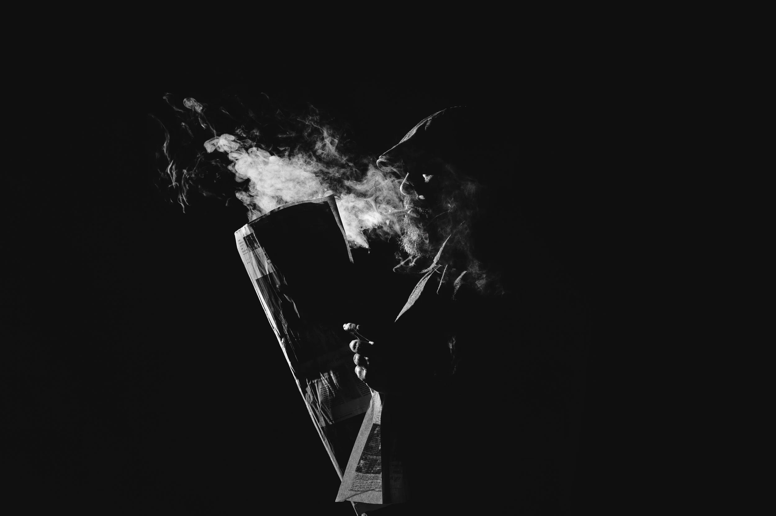 Black and white photograph of a person holding a lit cigarette, with smoke swirling around their face and hand, against a dark background.