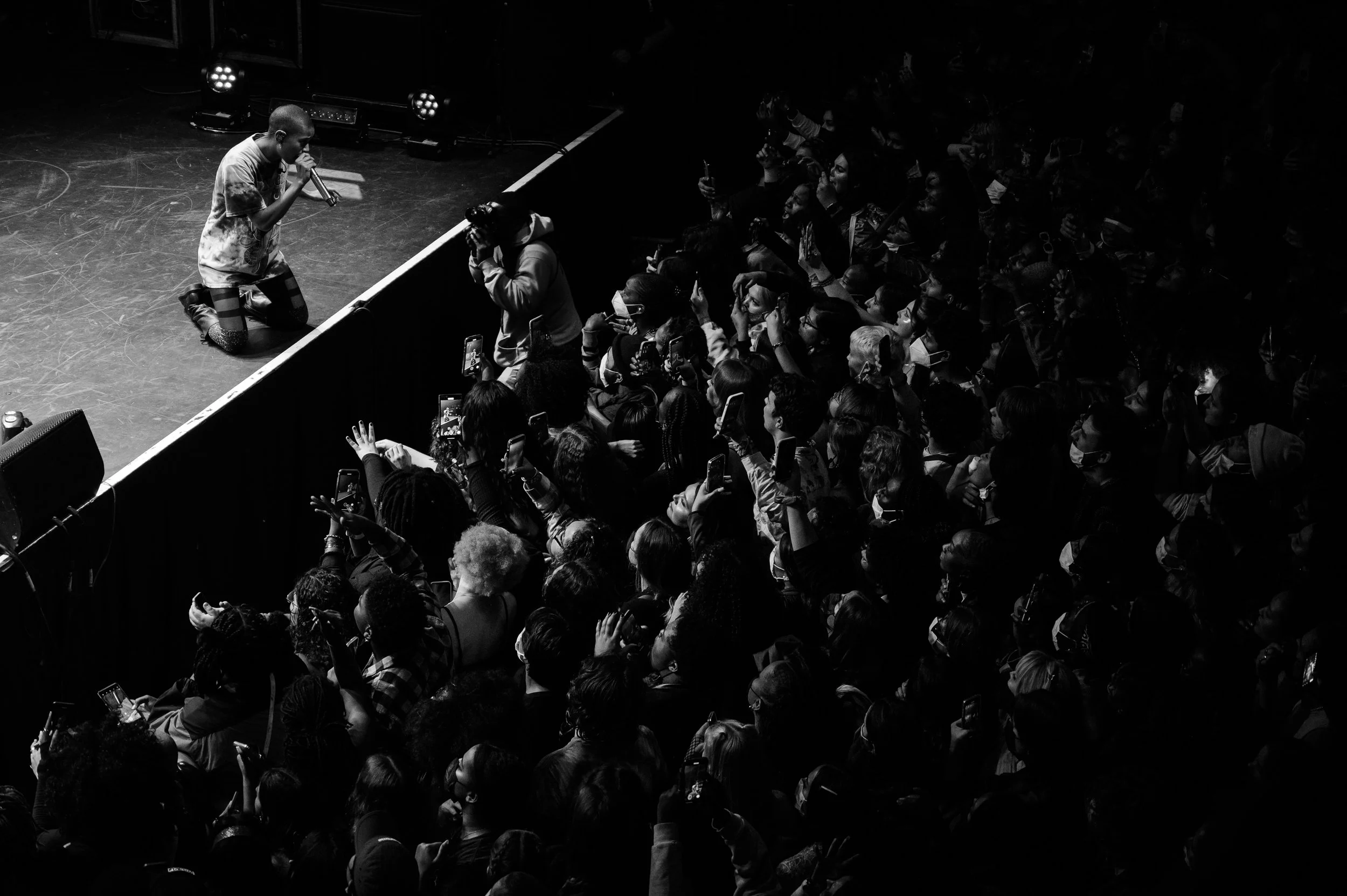 A performer kneeling on stage holding a microphone in front of a large crowd at a concert or event.