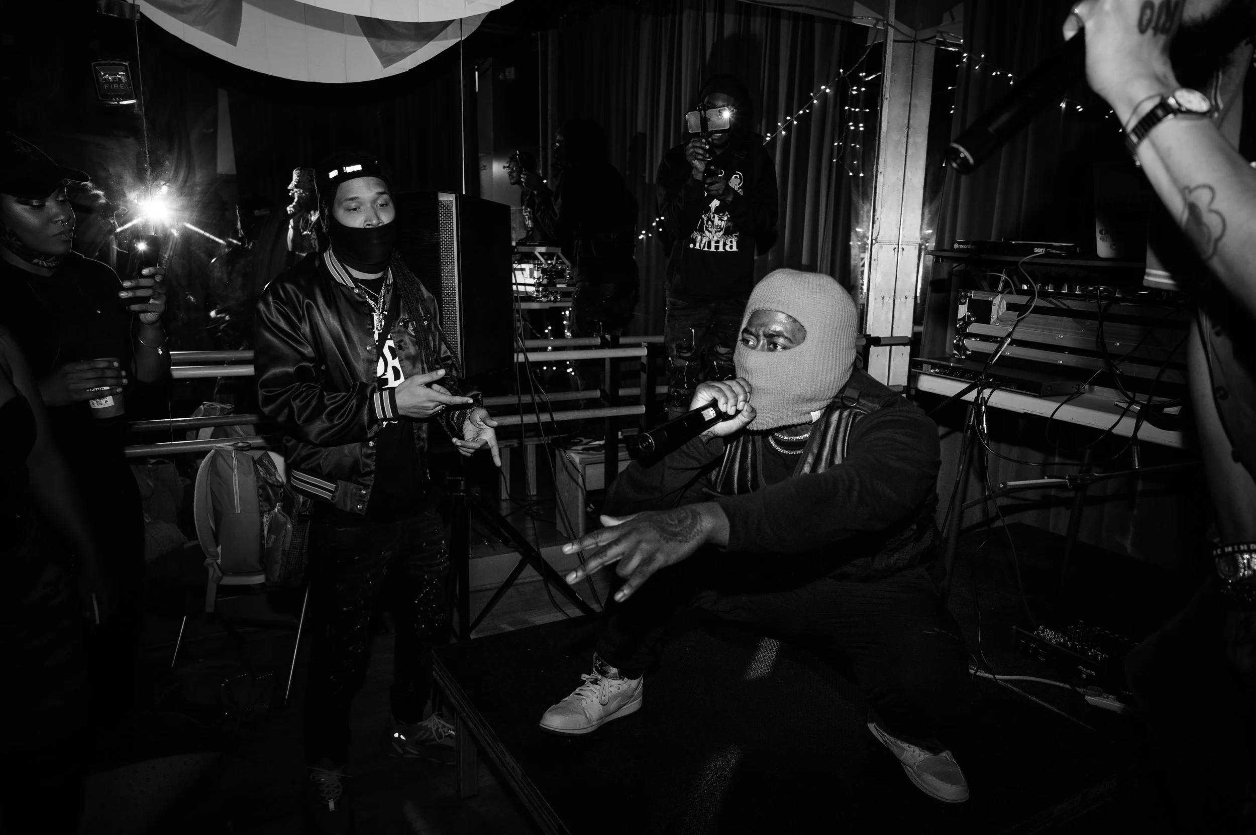 A group of people, including a singer with a beanie and mask, performing on stage with equipment and string lights in the background at a music event.
