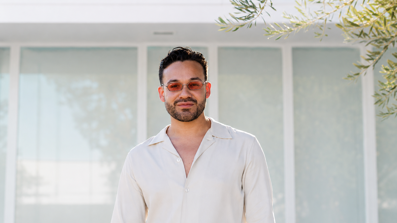 A man with dark hair, beard, and light skin wearing a cream-colored shirt and orange-tinted sunglasses standing outdoors in front of a modern glass building with greenery.