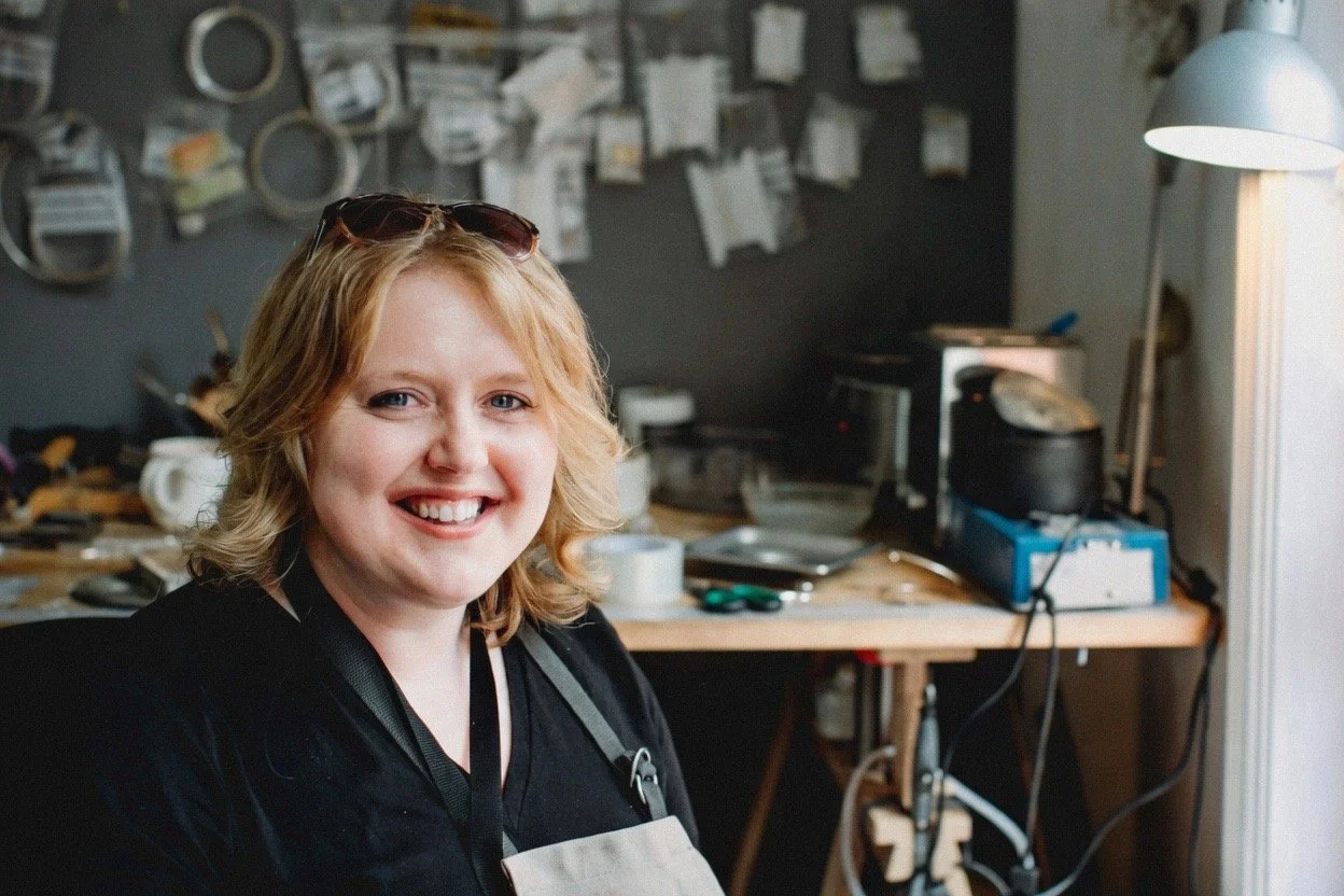 Woman with blonde hair smiling in a workshop or kitchen with tools and supplies in the background.