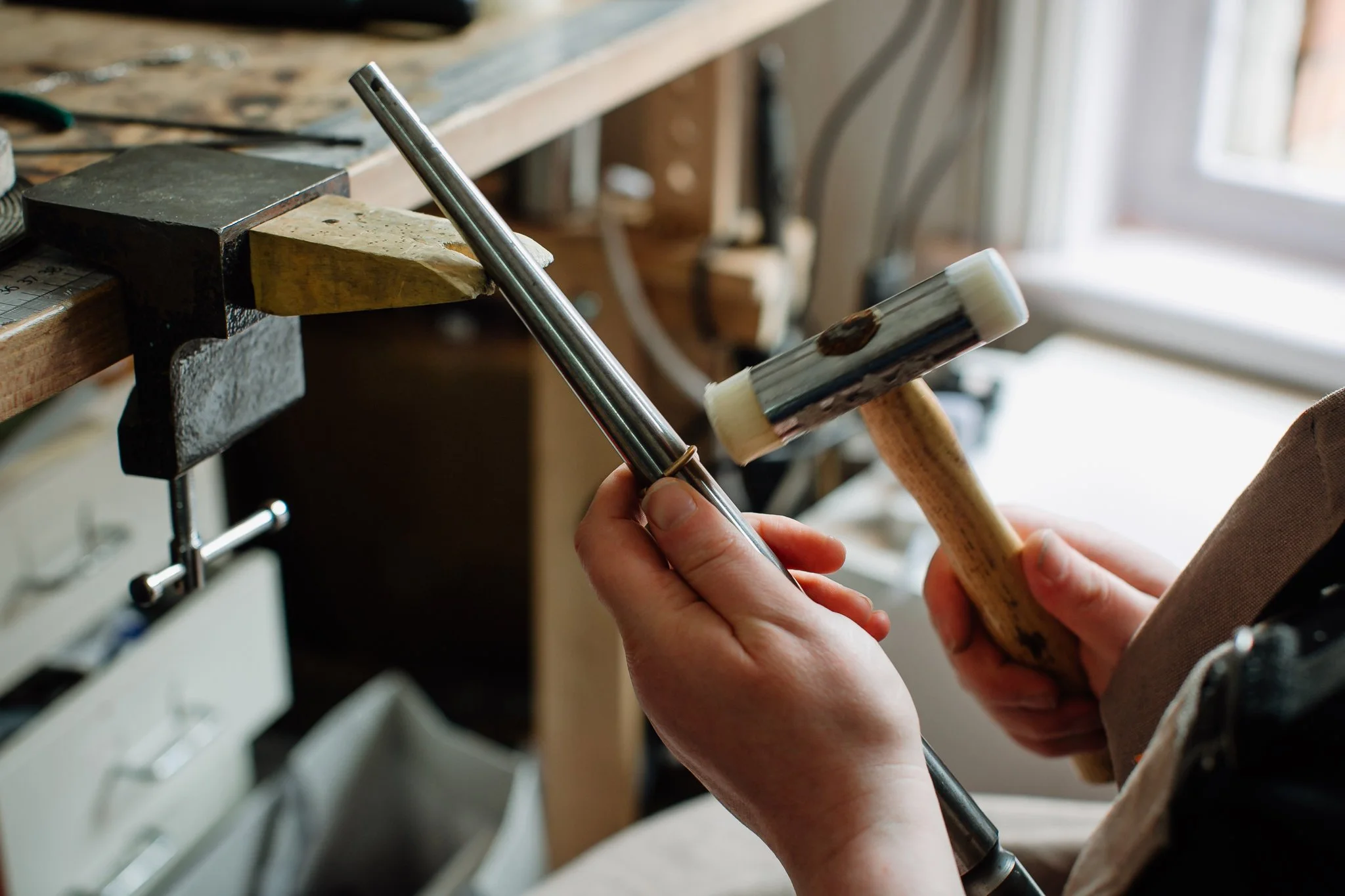 A person working in a workshop holding a mallet and a chisel near a piece of wood clamped to a workbench, with tools and workshop items in the background.