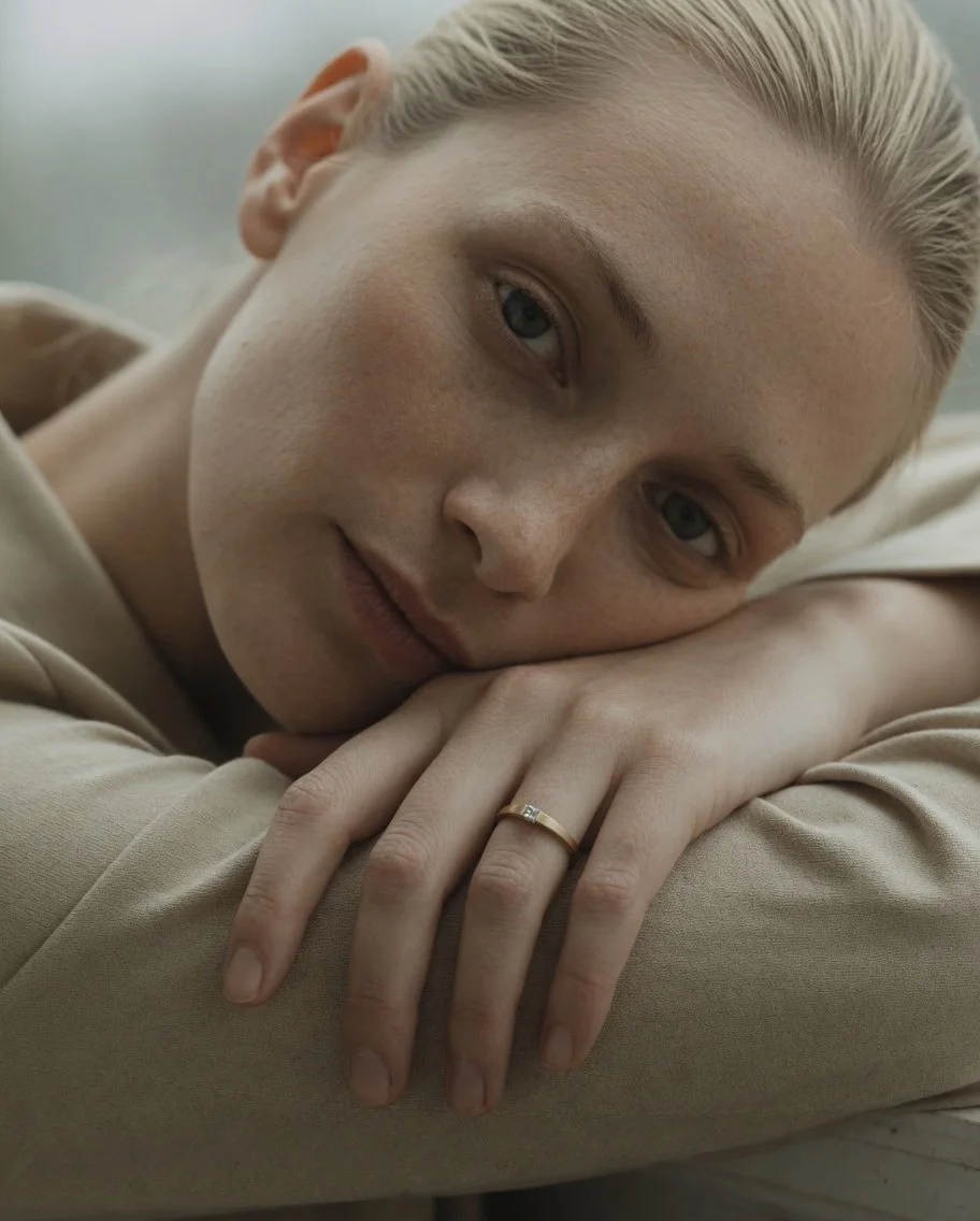 A young woman with blonde hair and light skin rests her head on her folded arms, gazing softly at the camera with a gentle expression.