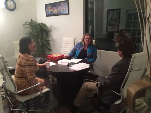 hree people sit around a conference table in an office, appearing to have a discussion with papers and coffee on the table.