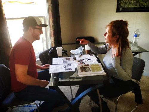 Two people sit at a small table indoors, discussing papers as one gestures while the other listens.