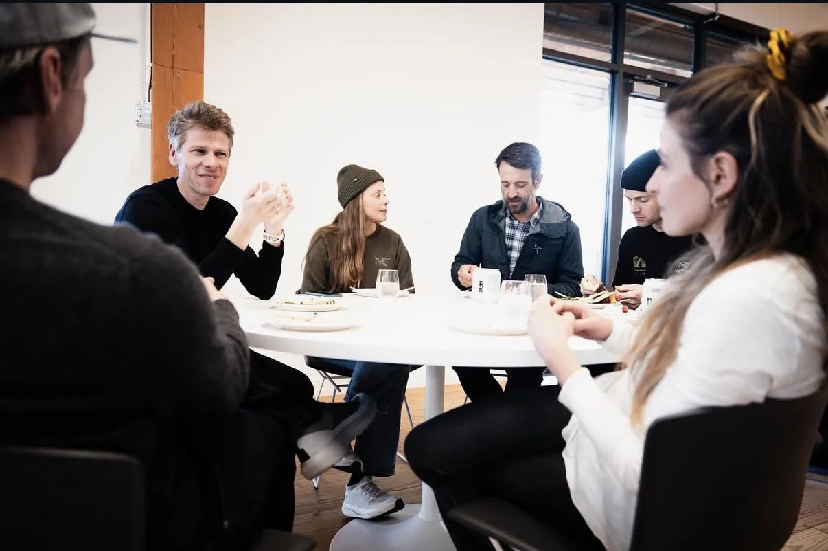 Group of six people sitting around a white round table in a casual meeting or discussion, with food and drinks on the table.