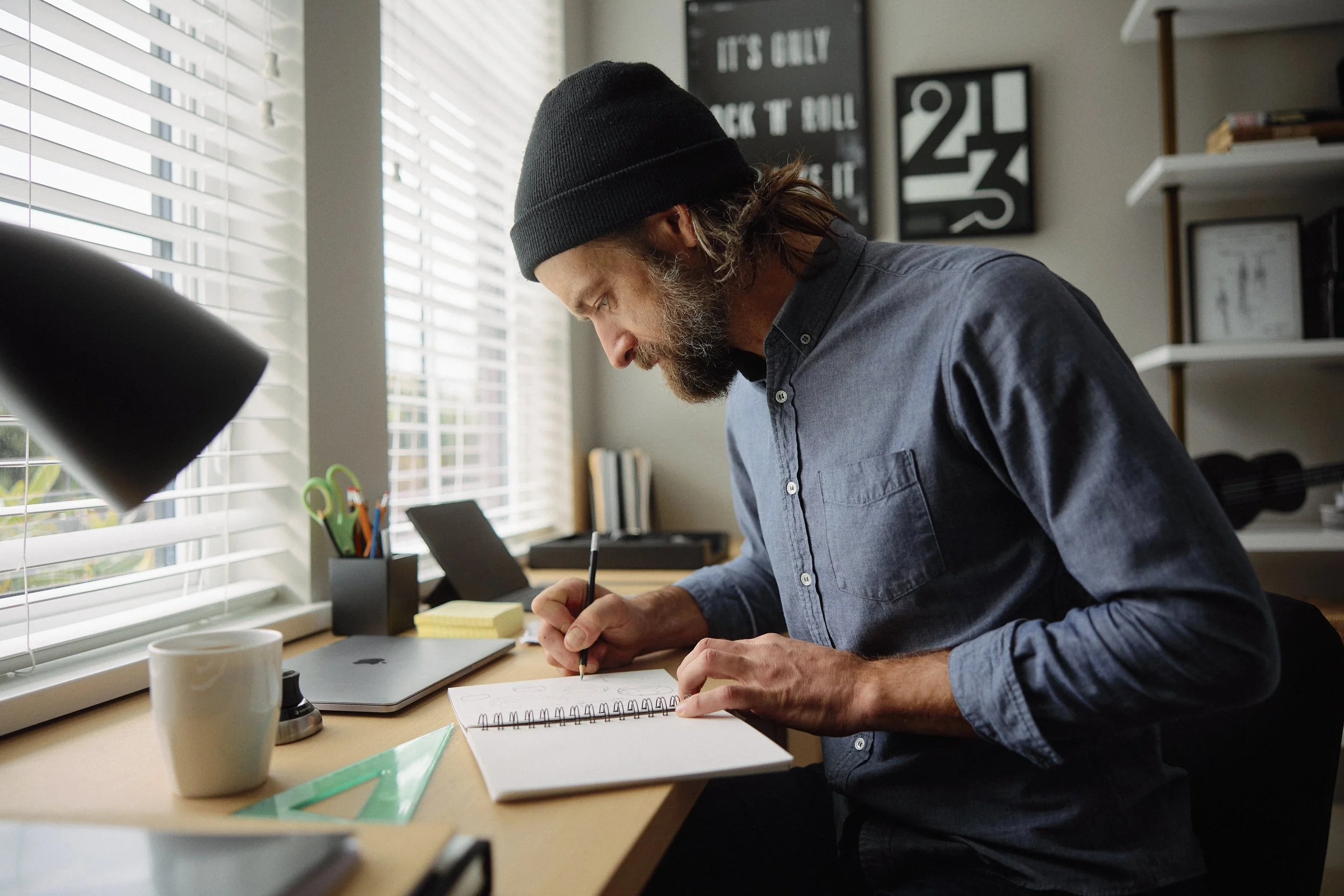 A man wearing a black beanie and blue shirt taking notes in a spiral notebook at a desk near a window.