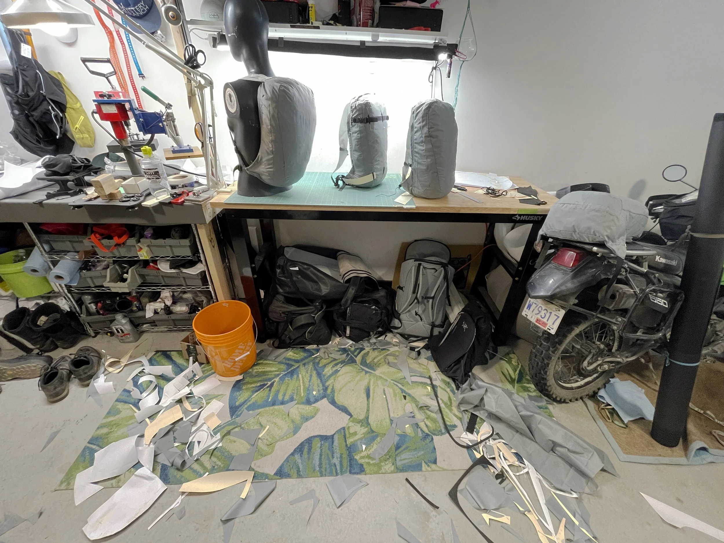 Workshop with various backpacks and gear, a motorcycle, and a cluttered workbench. Pieces of fabric or paper scraps on the floor and a colorful rug beneath.