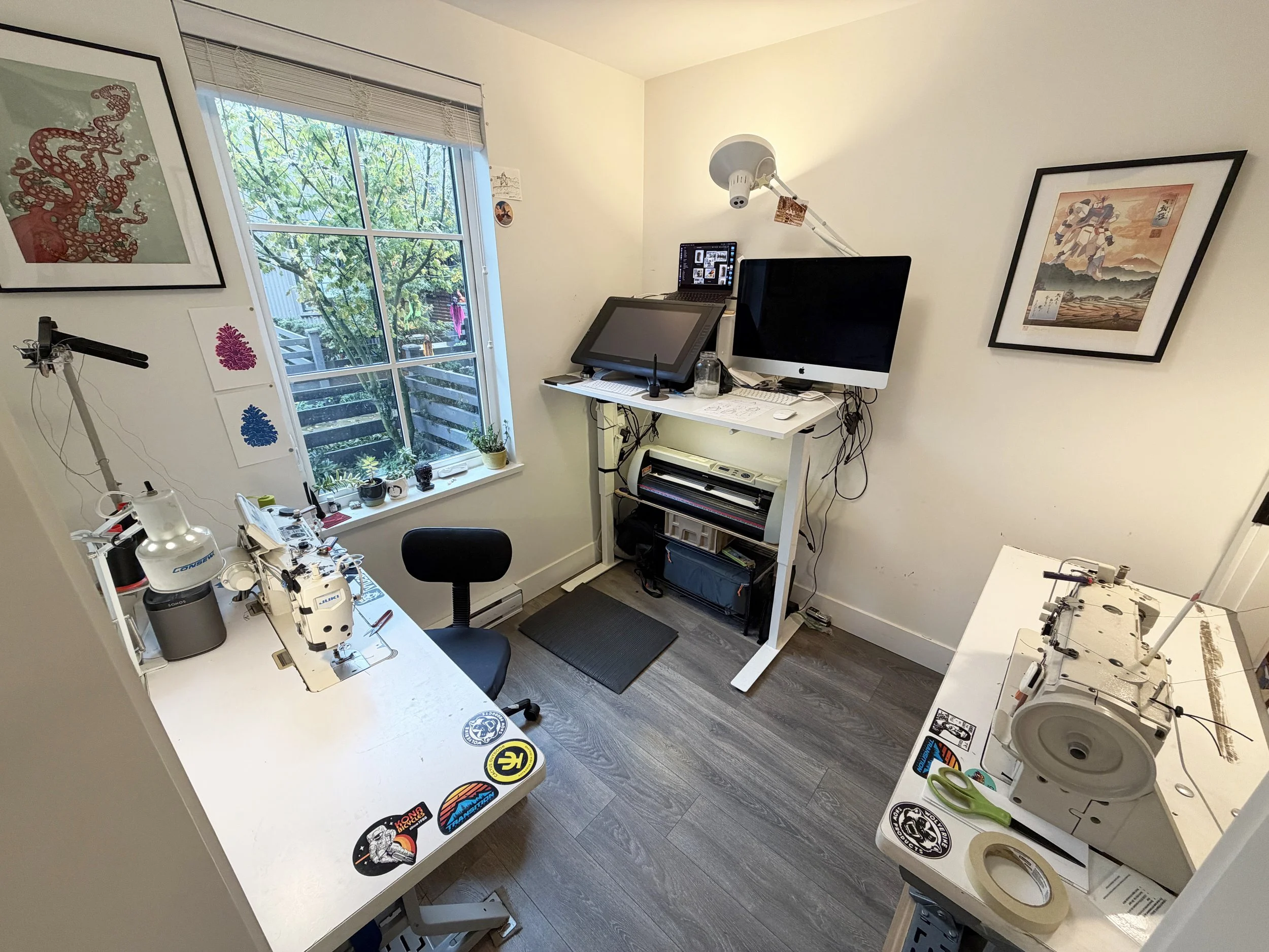 A small home workshop with a window showing greenery outside. There are two workstations with various tools and equipment, including a sewing or embroidery machine, a computer, and a drawing tablet, along with wall art, stickers, and potted plants.